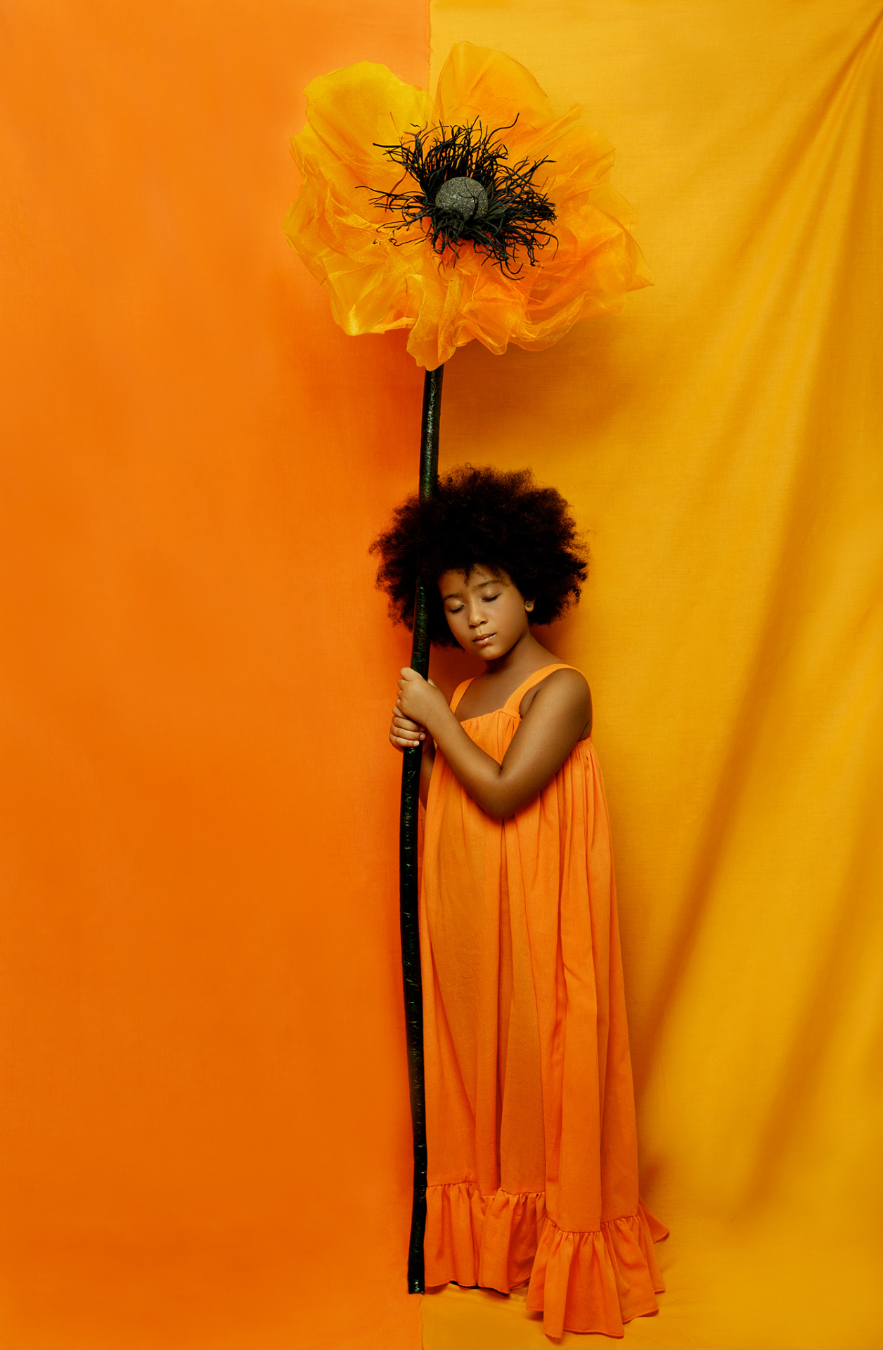 Ritratto poetico di bambina in abito arancione con fiore oversize e occhi chiusi, su sfondo bicolore, realizzato da Claudia Spaziani a Bologna.