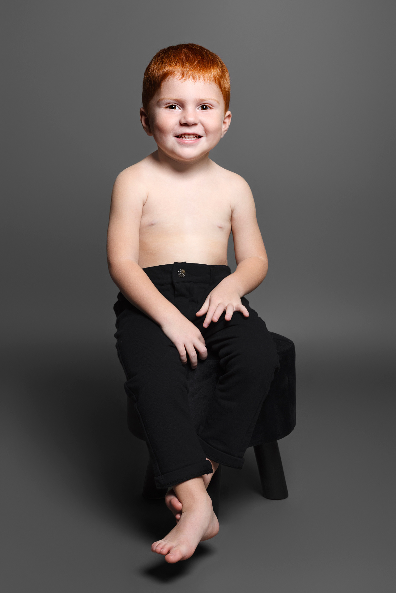 Ritratto minimalista di bambino dai capelli rossi, seduto su sgabello nero con pantaloni scuri, realizzato da Claudia Spaziani a Bologna.