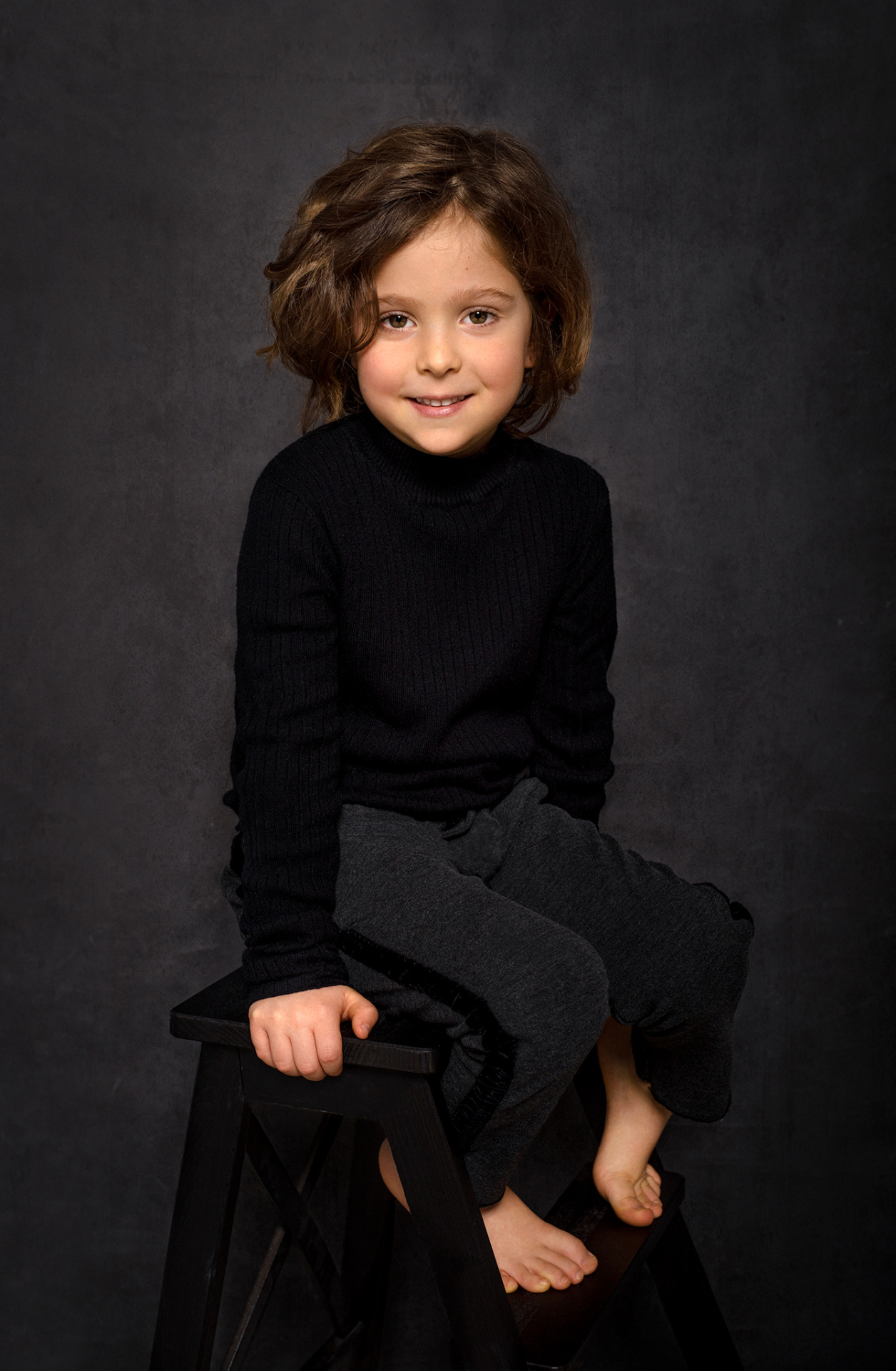 Ritratto in studio di bambino a piedi nudi con maglia nera e pantaloni grigi, su sfondo scuro, realizzato da Claudia Spaziani a Bologna.