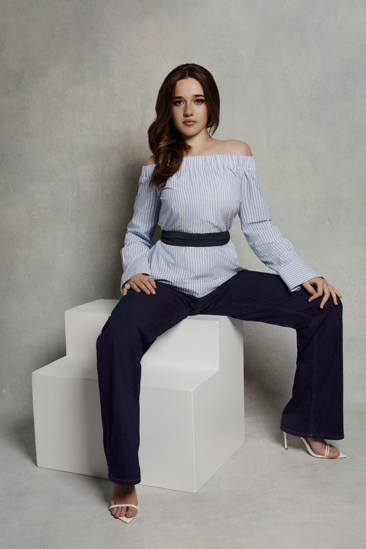 Ritratto contemporaneo di adolescente in camicia a righe e pantaloni blu, seduta su blocco geometrico, scattato da Claudia Spaziani nel suo studio fotografico a Bologna.