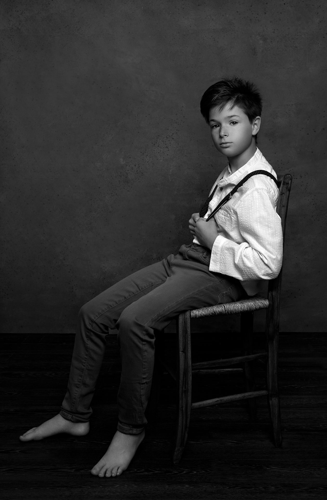 Ritratto monocromatico di bambino con camicia bianca e bretelle, seduto su sedia in legno, realizzato da Claudia Spaziani nel suo studio fotografico a Bologna.