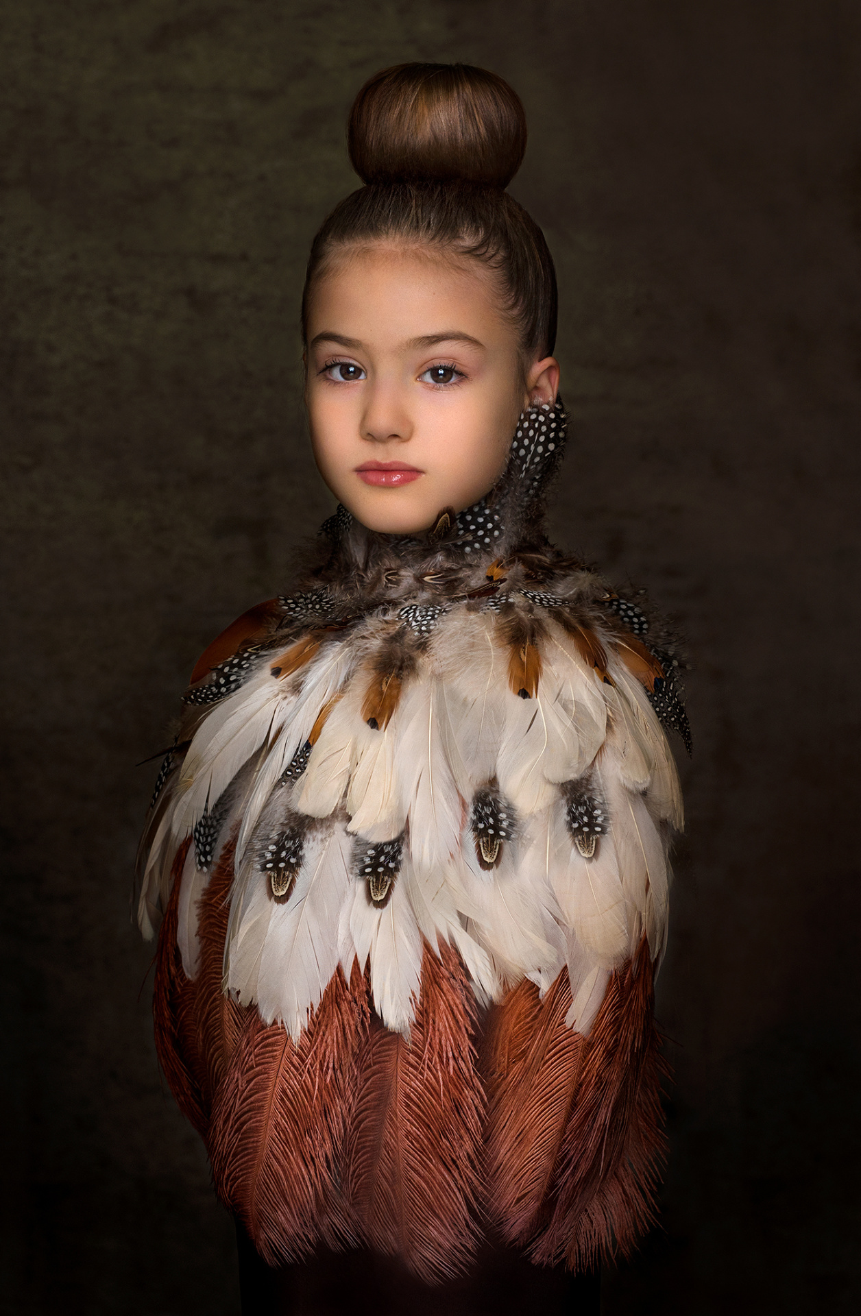 Ritratto artistico di bambina con abito composto da piume multicolore e chignon alto, su sfondo scuro, realizzato da Claudia Spaziani nel suo studio fotografico a Bologna.