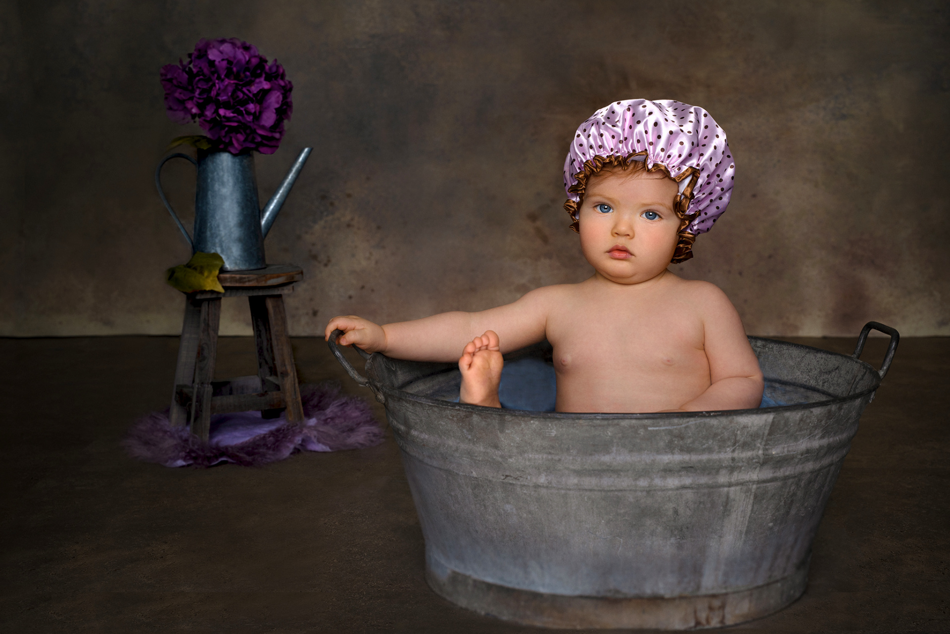 Foto creativa di bambina in posa dentro un catino di zinco, con cuffia rosa a pois e fiori viola, scattata da Claudia Spaziani nel suo studio a Bologna.