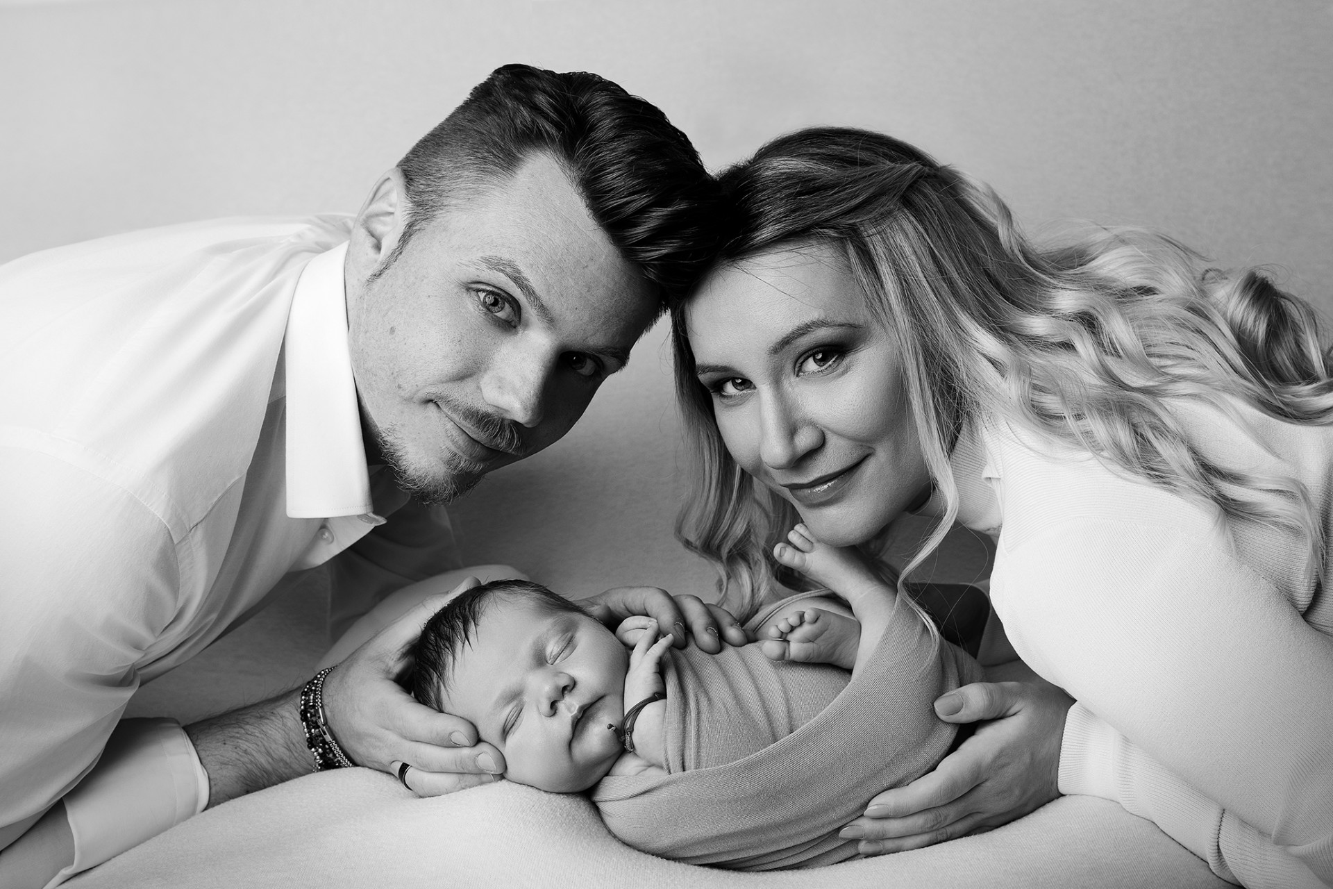 Fotografia in bianco e nero di genitori che cullano neonato maschio avvolto in coperta, scattata da Claudia Spaziani fotografa a Bologna. Immagine intima che celebra amore e legame familiare.