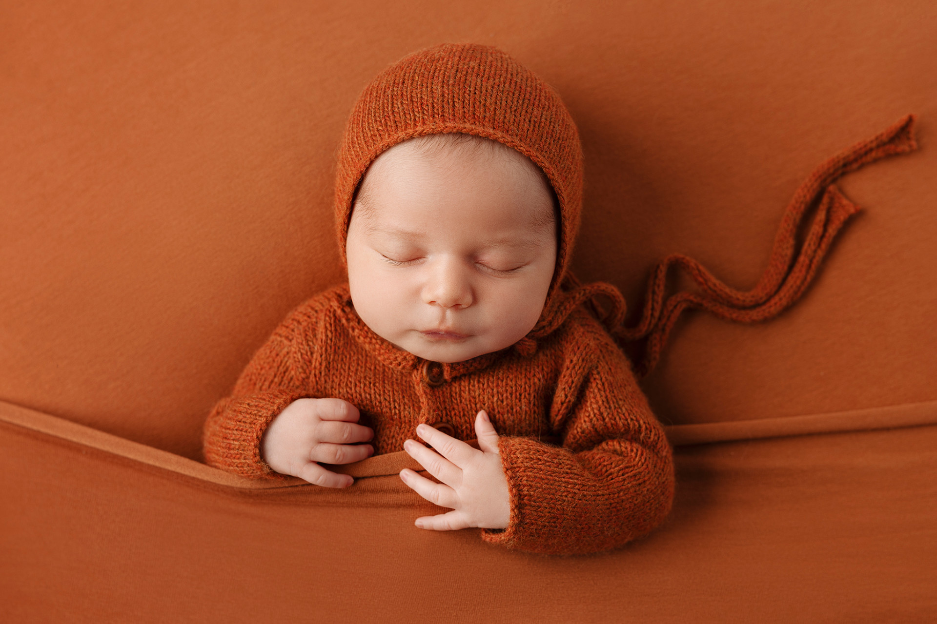 Fotografia neonatale a Bologna di Claudia Spaziani: neonata addormentata in abito e cuffietta color ruggine, posata su tessuto coordinato. Immagine calda e monocromatica che trasmette comfort e tenerezza.
