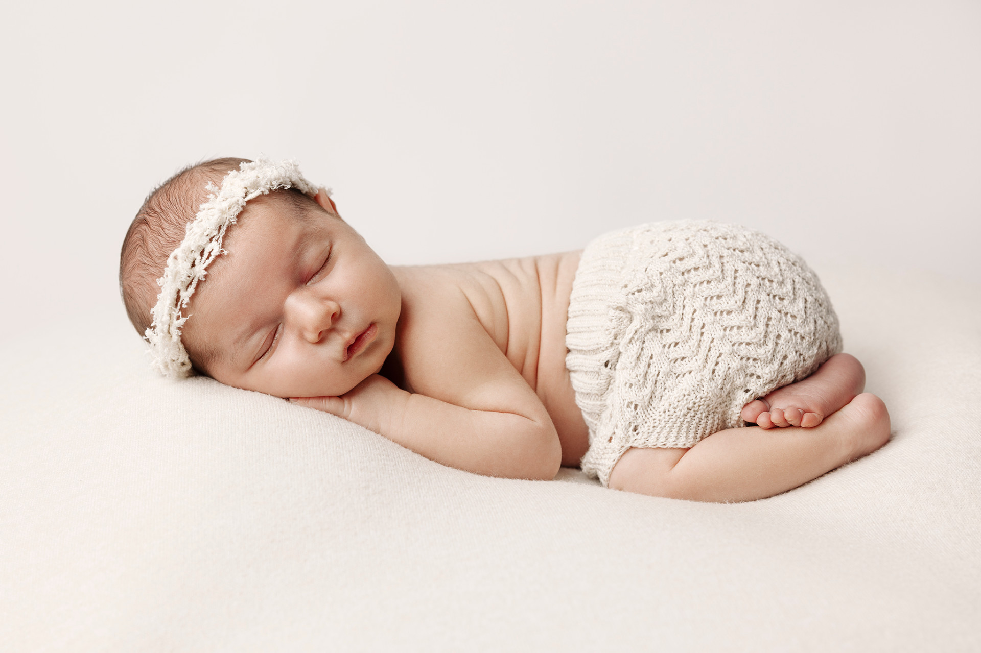 Fotografia neonatale a Bologna di Claudia Spaziani: neonata addormentata in outfit bianco con fascia floreale e pantaloncini lavorati a maglia. Immagine che esalta serenità e dolcezza.