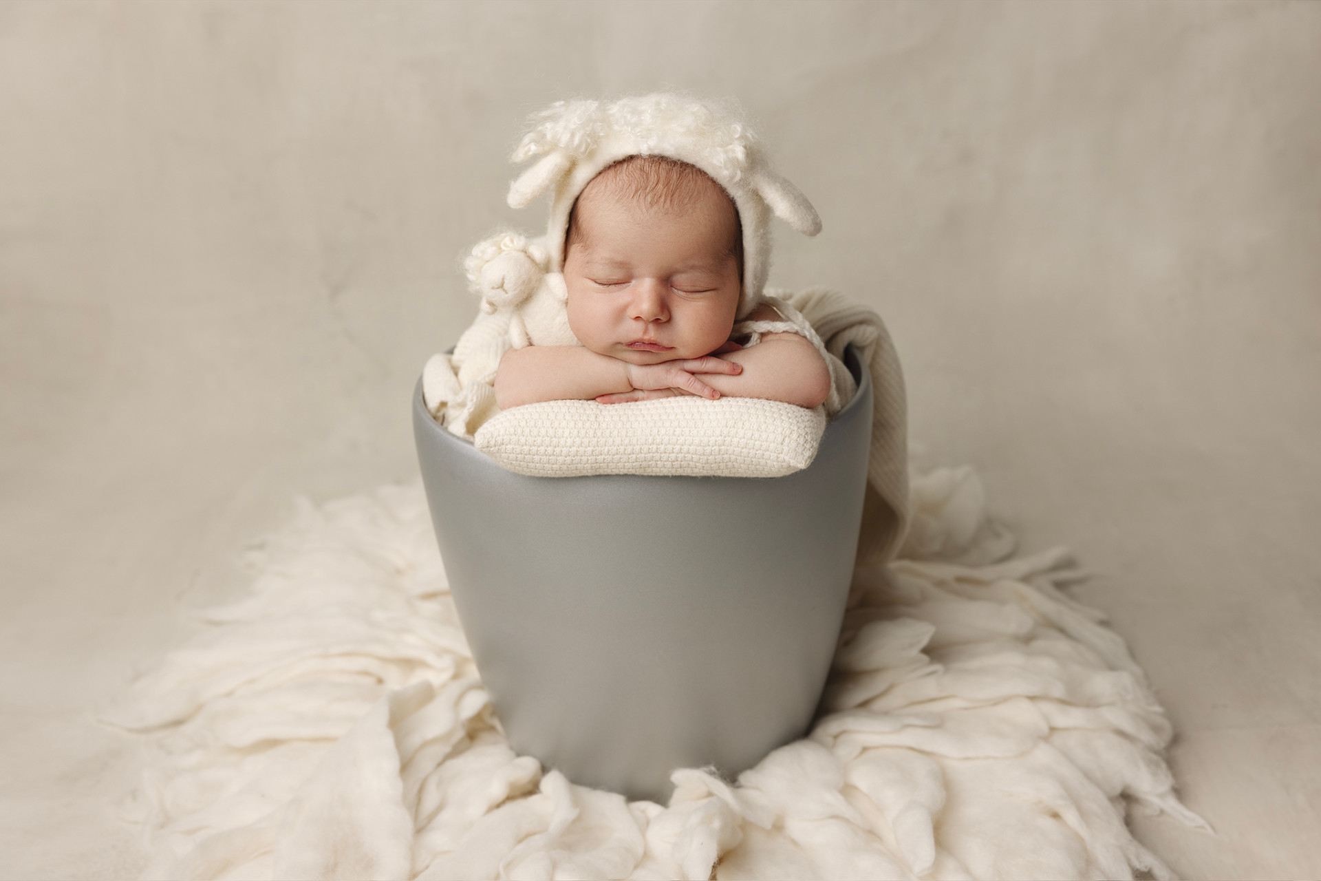 Foto creativa di neonata addormentata in outfit bianco con cappellino da agnellino, posata in secchiello grigio su imbottitura soffice, scattata da Claudia Spaziani a Bologna. Immagine dolce e fiabesca.