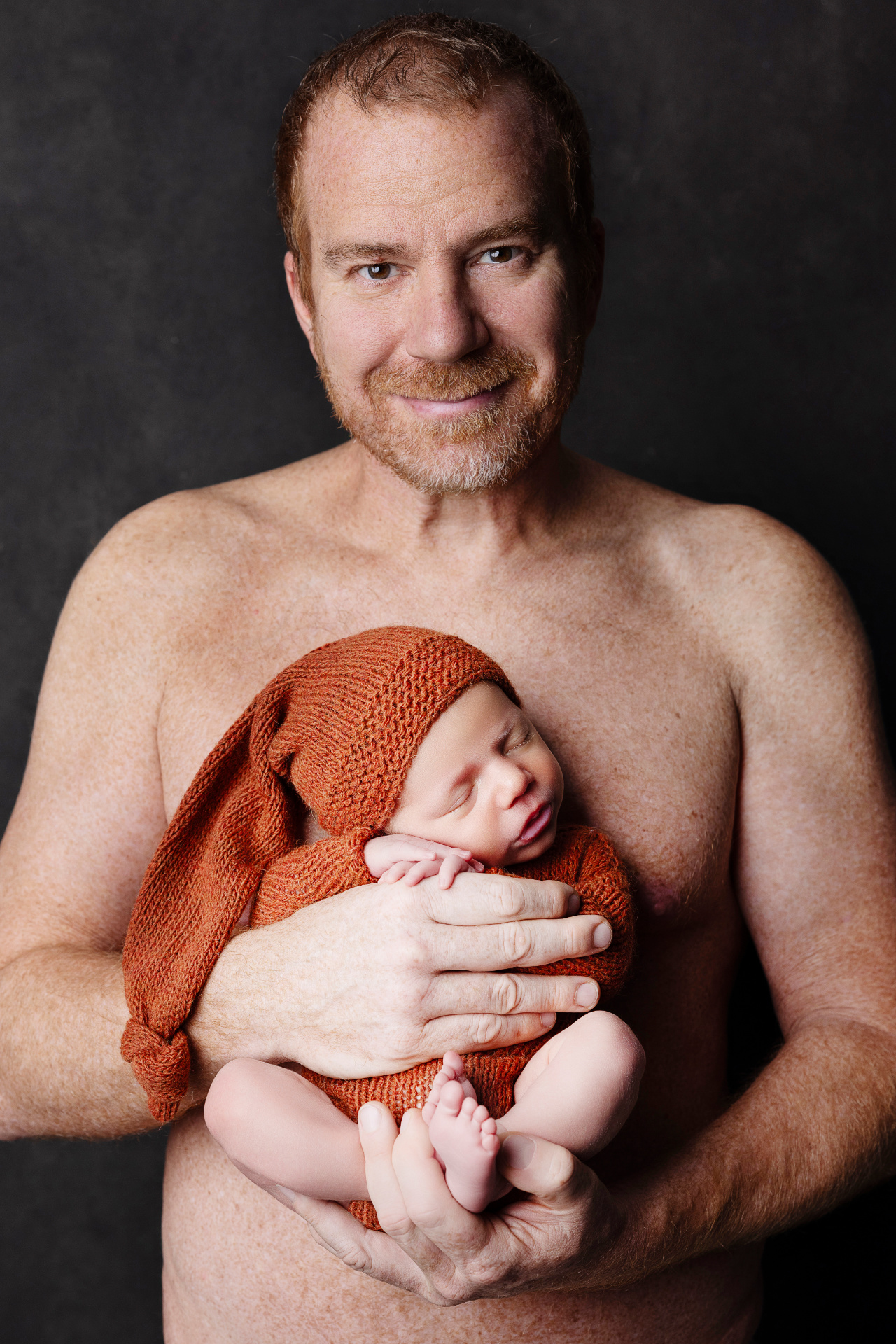 Ritratto di padre che tiene in braccio neonato maschio in abito marrone, fotografato nello studio di Claudia Spaziani a Bologna. Immagine che celebra amore e protezione.