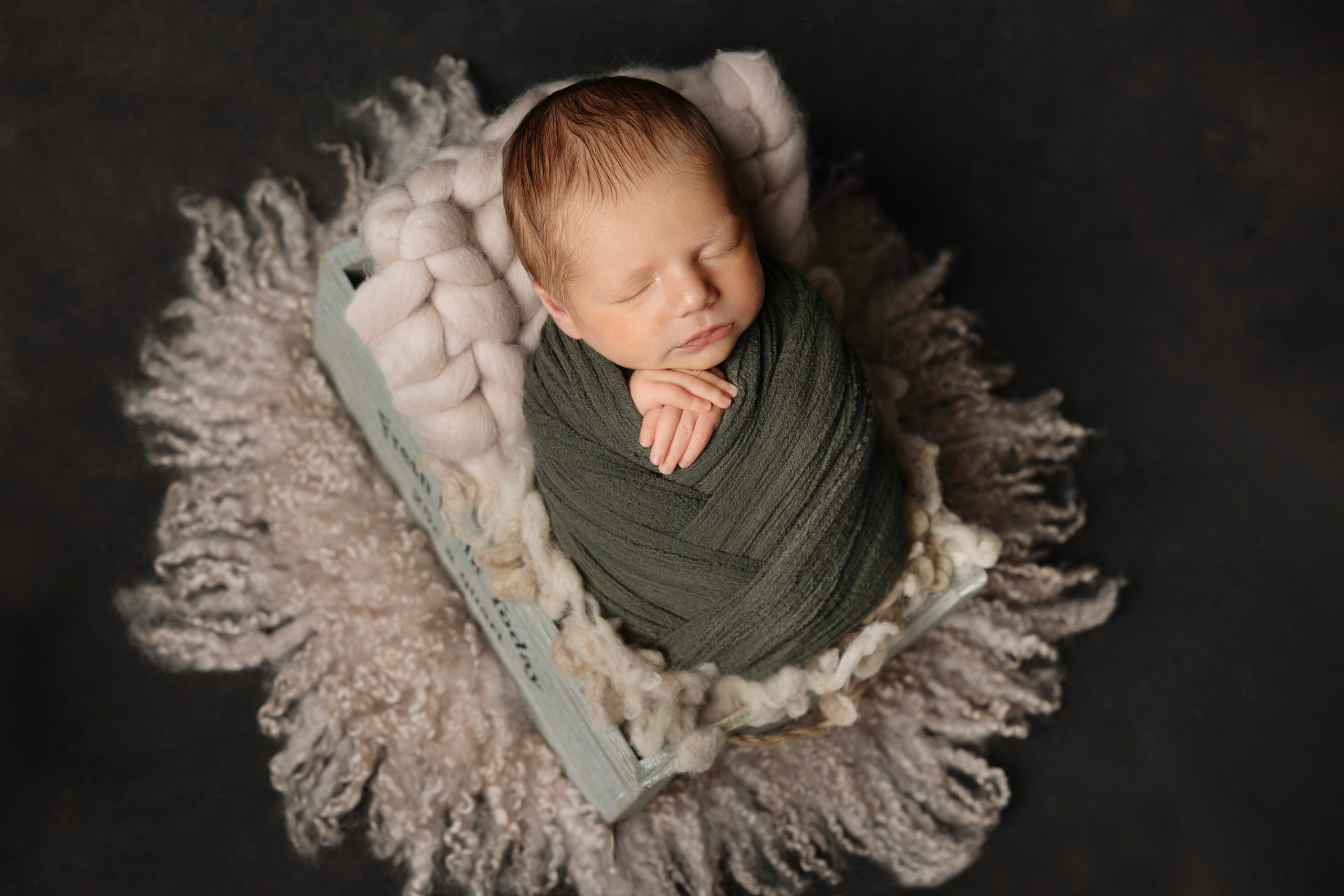 Ritratto di neonato maschio addormentato in fascia verde, posato in cassetta di legno azzurra con imbottitura soffice, fotografato nello studio di Claudia Spaziani a Bologna. Immagine calda e rassicurante che esalta innocenza e serenità.