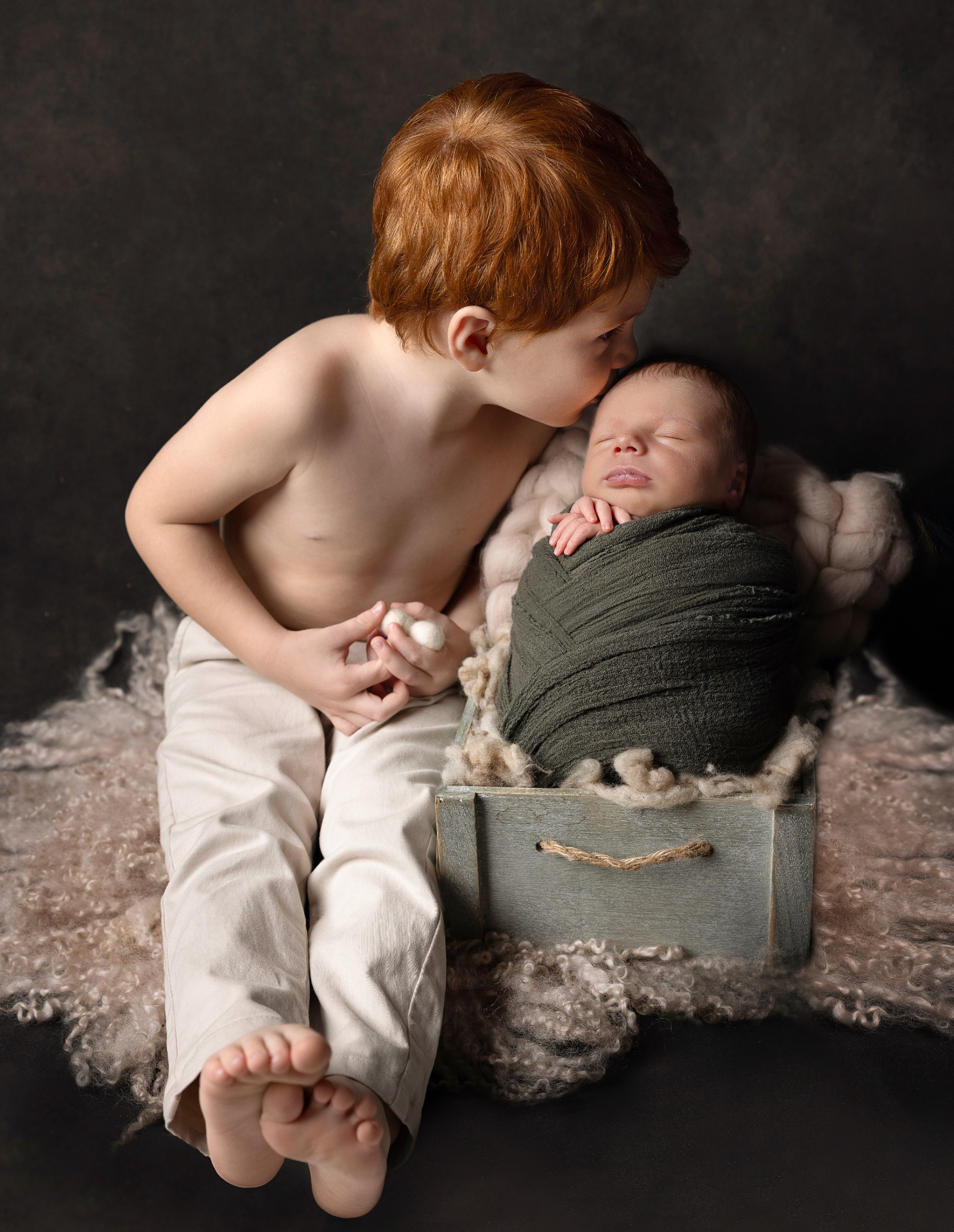 Fotografia di fratello maggiore che bacia neonato maschio avvolto in fascia verde, posato in cassetta di legno, scattata nello studio di Claudia Spaziani a Bologna. Immagine tenera e simbolica.