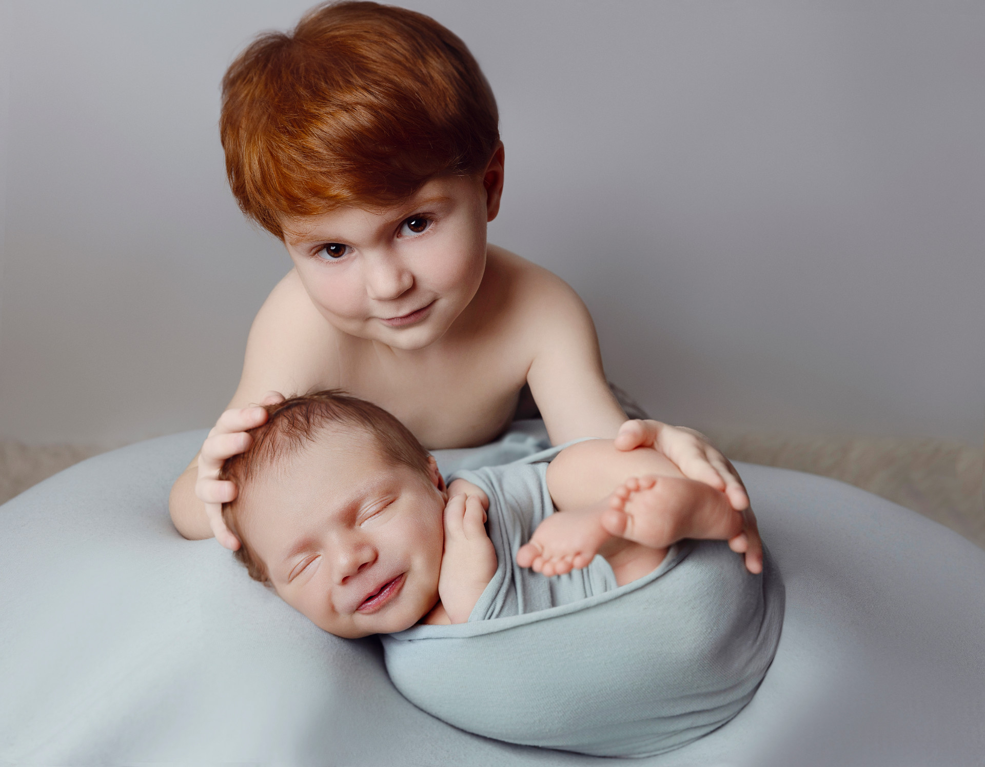 Fotografia di fratello maggiore che culla neonato maschio avvolto in fascia azzurra, scattata nello studio di Claudia Spaziani a Bologna. Immagine tenera che celebra il legame tra fratelli.