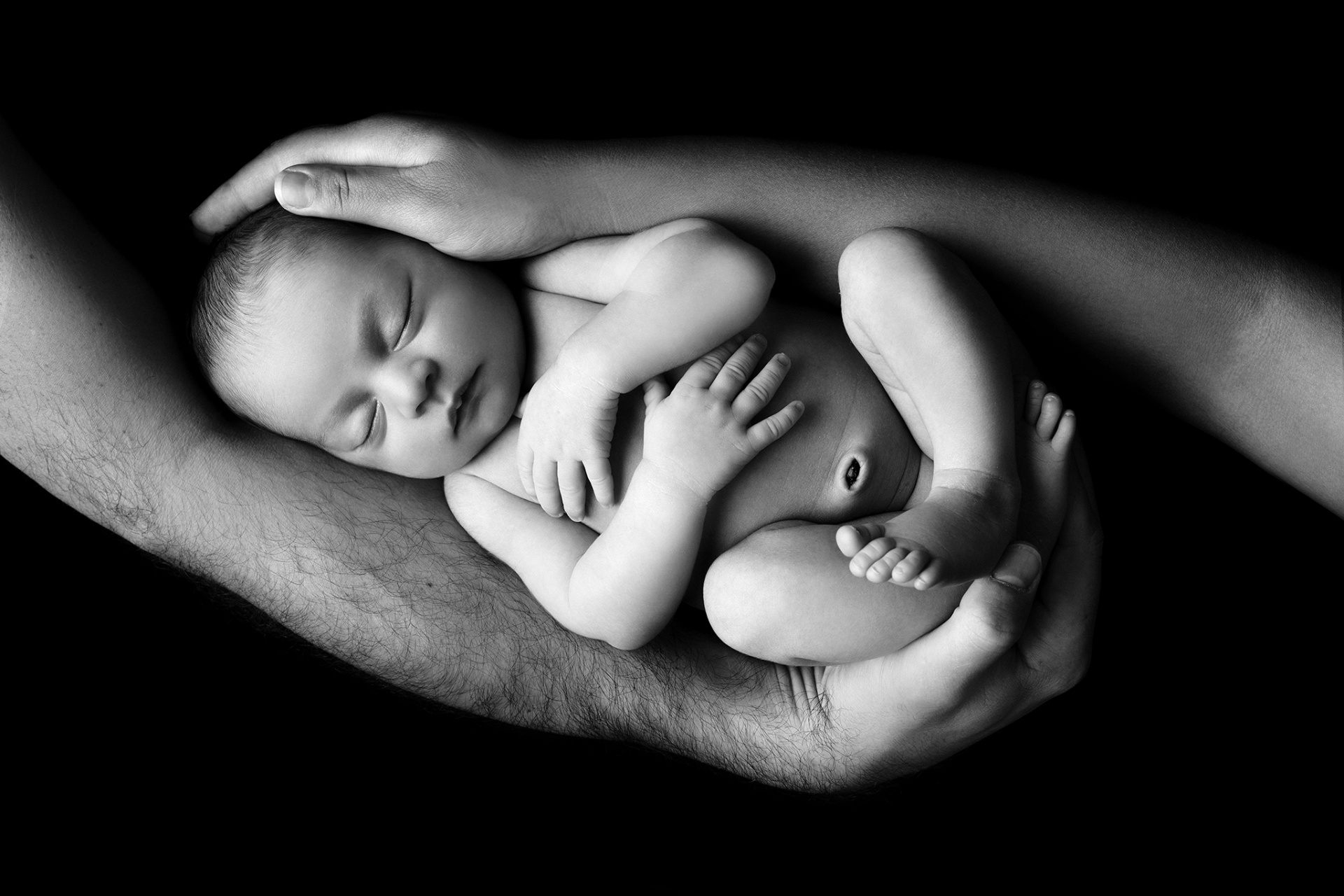 Ritratto in bianco e nero di neonata addormentata in posizione fetale, sorretta da mani adulte, fotografata nello studio di Claudia Spaziani a Bologna. Immagine intima e suggestiva.