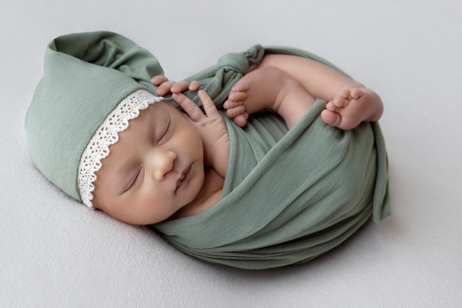 Foto artistica di neonata addormentata in fascia verde con cappellino coordinato e bordo in pizzo, scattata nello studio di Claudia Spaziani a Bologna. Atmosfera serena e delicata.