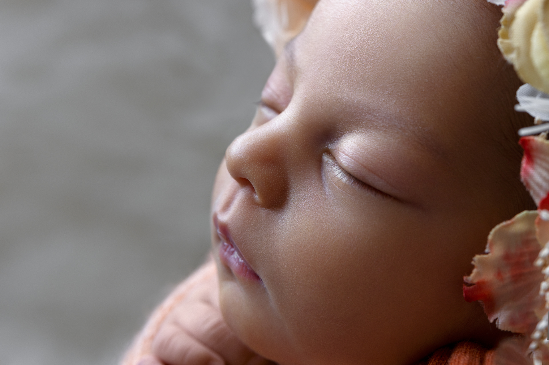 Primo piano di neonata addormentata con fascia pesca e cerchietto floreale rosso, bianco e giallo, realizzato da Claudia Spaziani fotografa a Bologna. Immagine intima e tenera.