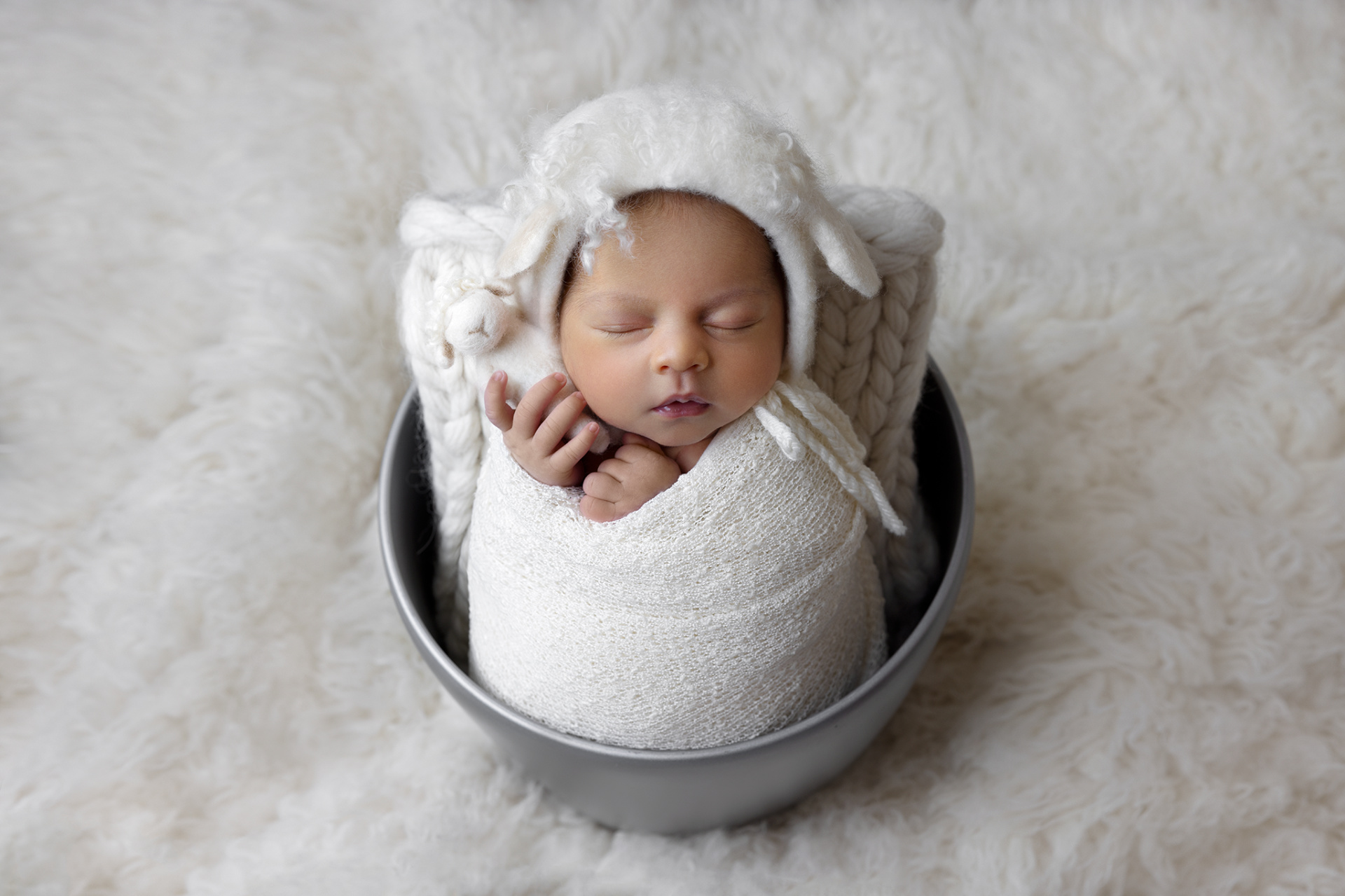 Fotografia neonatale a Bologna di Claudia Spaziani: neonata addormentata in coperta bianca, con cappellino a orecchie di agnello, posata in ciotola grigia su sfondo soffice. Immagine dolce e fiabesca