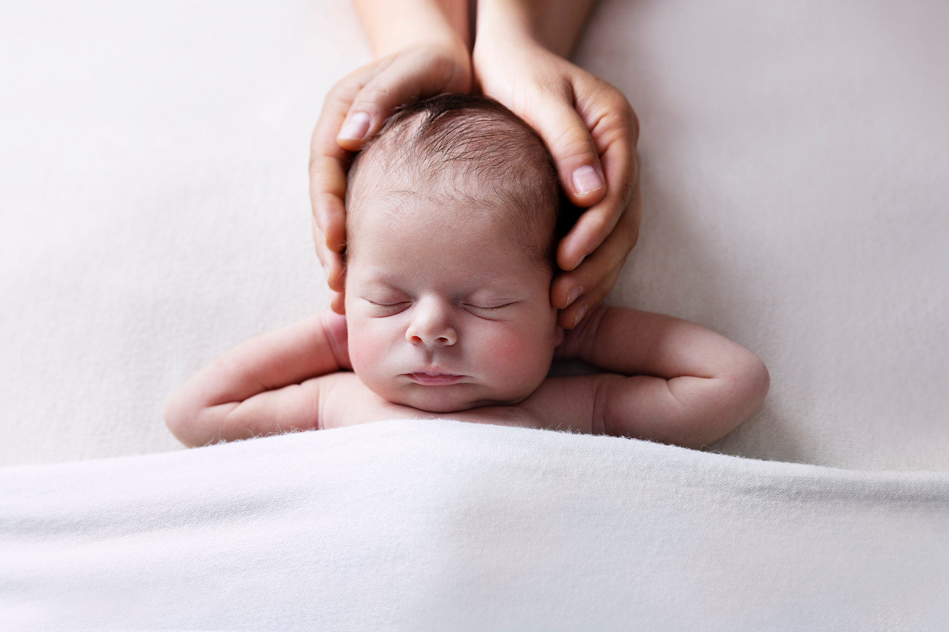 Fotografia neonatale a Bologna di Claudia Spaziani: neonato addormentato su superficie bianca, coperto da coperta soffice, con mani adulte che sostengono delicatamente la testa, simbolo di amore e protezione