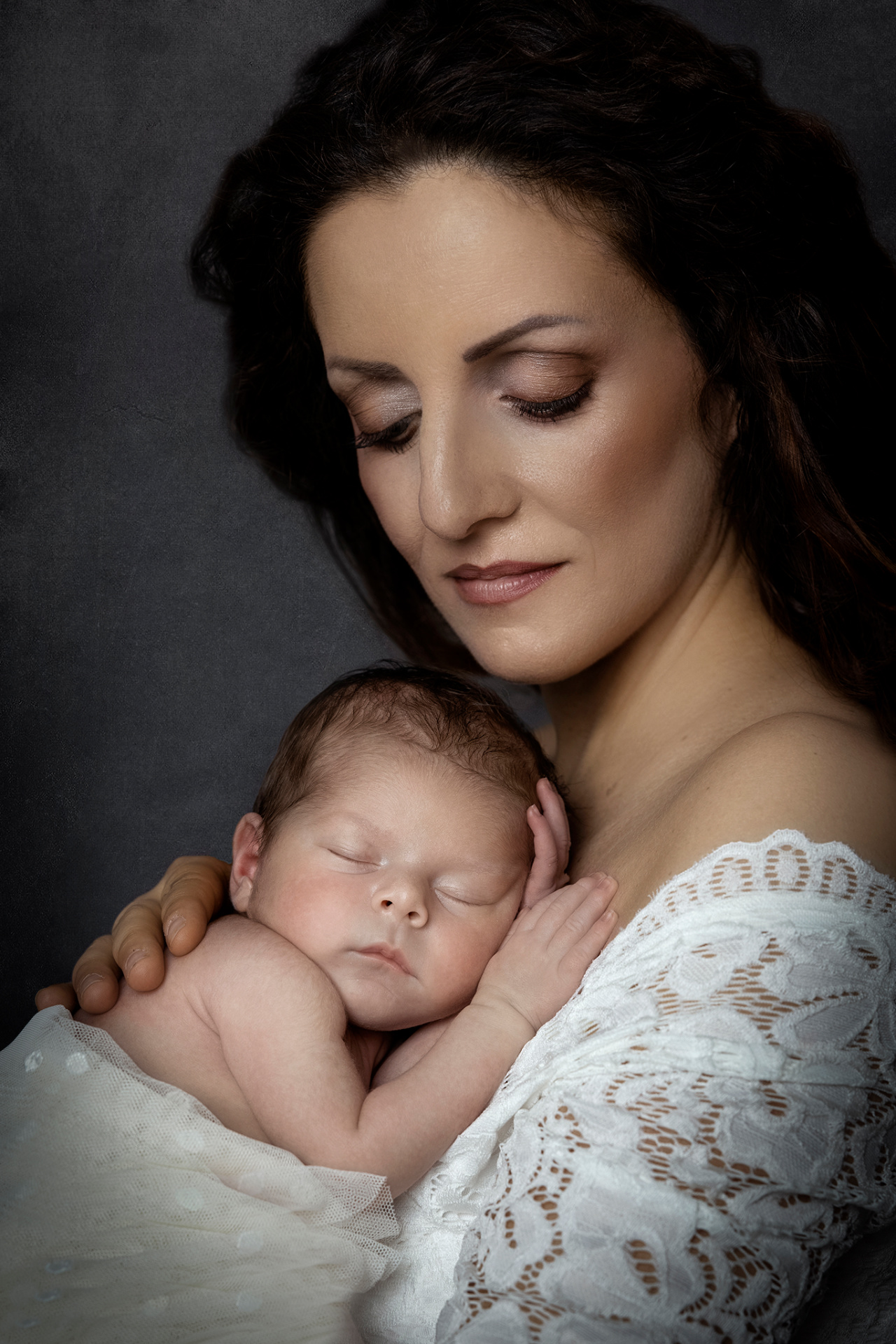 Ritratto intimo di madre con neonato addormentato, realizzato da Claudia Spaziani fotografa a Bologna. La mamma in abito di pizzo bianco culla il bambino con dolcezza, in un’atmosfera elegante e raccolta.