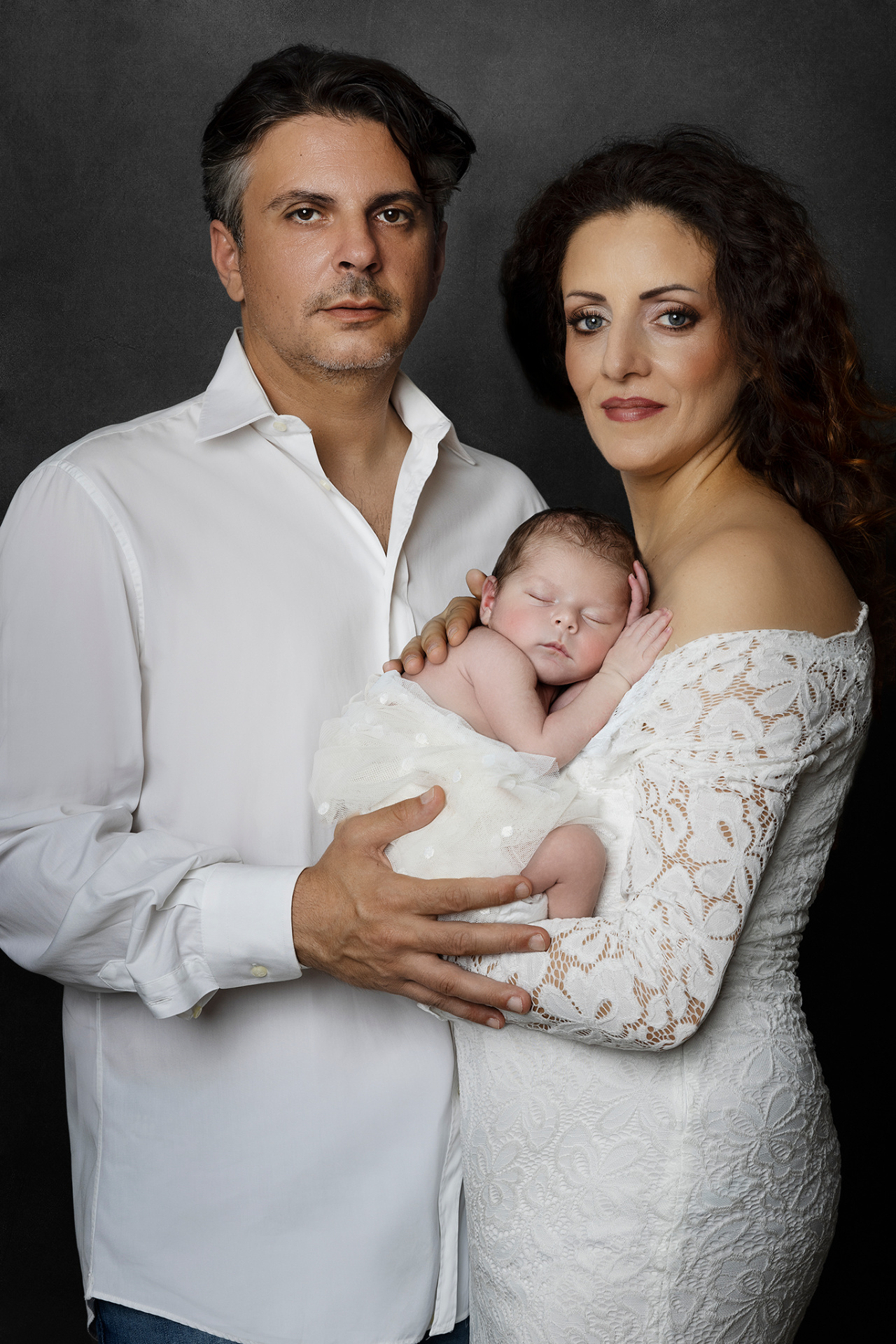 Foto di famiglia con neonato addormentato, realizzata da Claudia Spaziani fotografa a Bologna. Genitori in abiti bianchi sostengono il bambino con tenerezza, in un ritratto elegante e senza tempo