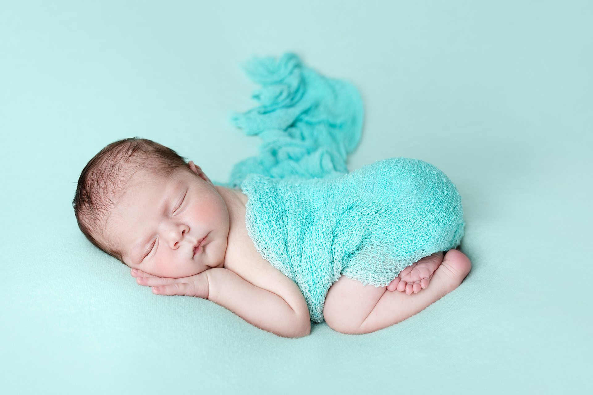 Foto artistica di neonato addormentato su superficie turchese, avvolto in fascia coordinata, scattata nello studio fotografico di Claudia Spaziani a Bologna. Immagine che trasmette calma e tenerezza.