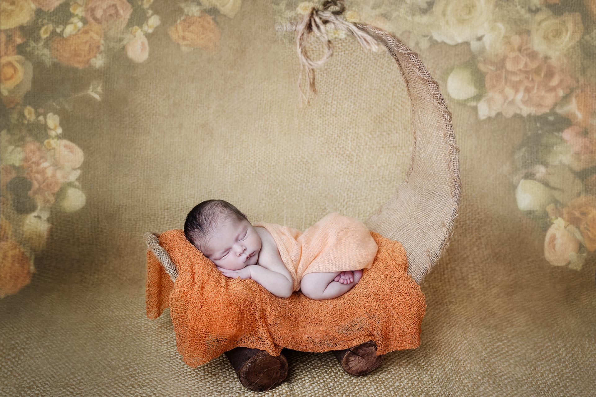 Ritratto creativo di neonato avvolto in fascia arancione, posato su culla a forma di luna nello studio fotografico di Claudia Spaziani a Bologna. Ambientazione calda e fiabesca che celebra la nascita.