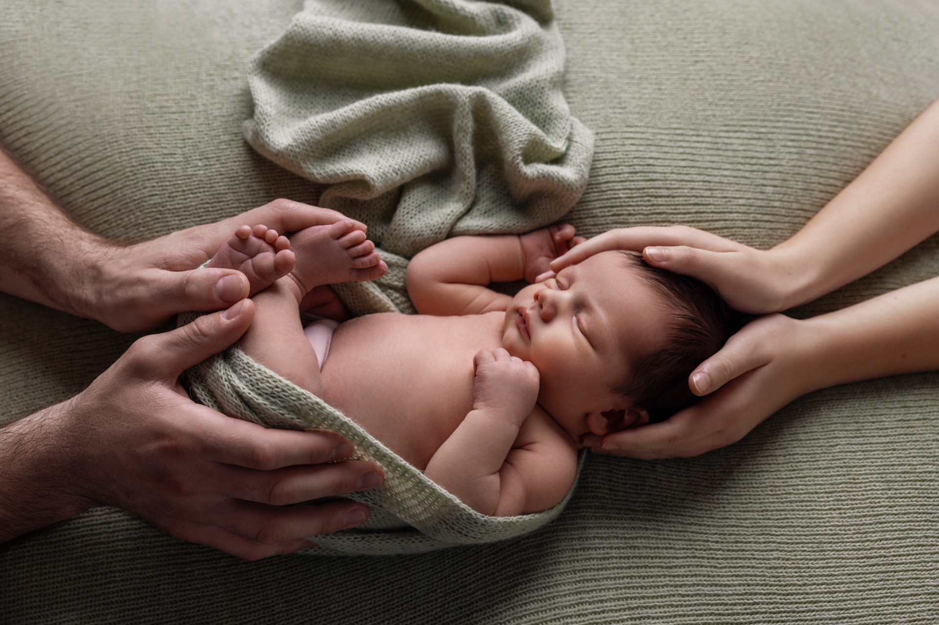 Servizio newborn a Bologna di Claudia Spaziani: neonato che dorme su coperta verde, sorretto con delicatezza da mani adulte.
