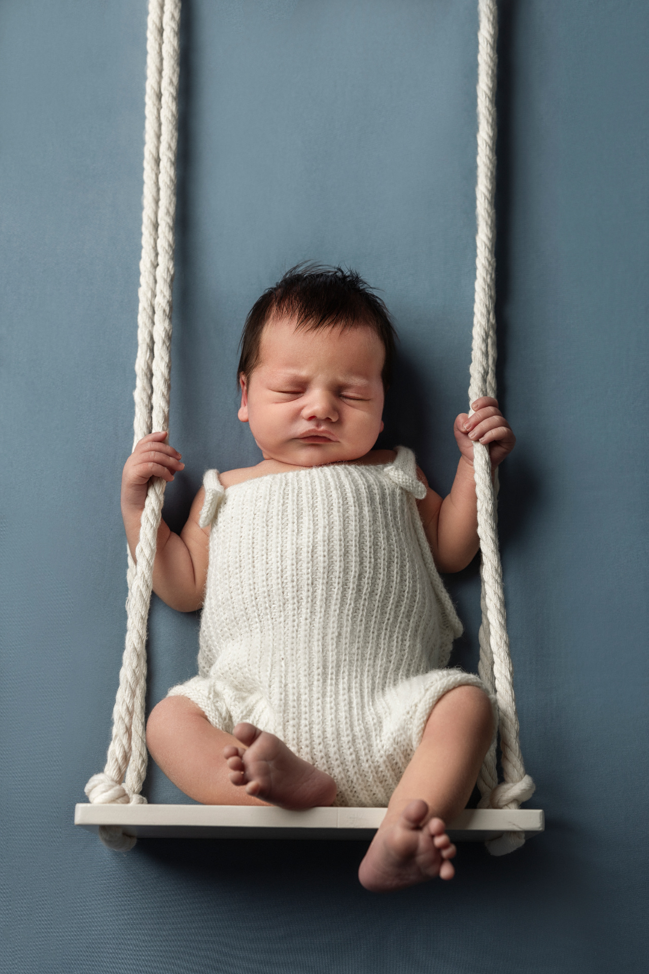Claudia Spaziani fotografa newborn a Bologna: neonato che riposa su altalena bianca con sfondo blu, in posa dolce e serena.