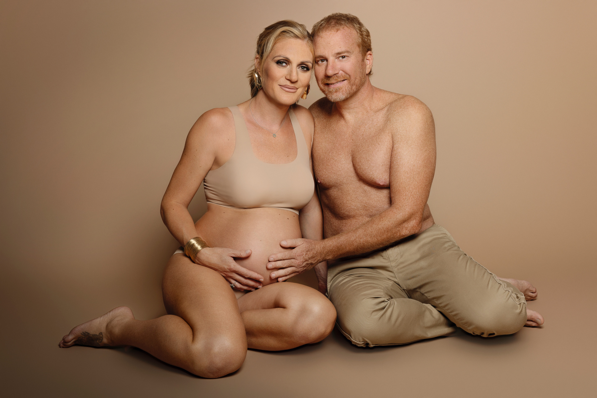 Servizio fotografico maternità a Bologna con Claudia Spaziani: coppia seduta su sfondo beige, donna incinta in bikini beige e gioielli dorati, posa affettuosa.