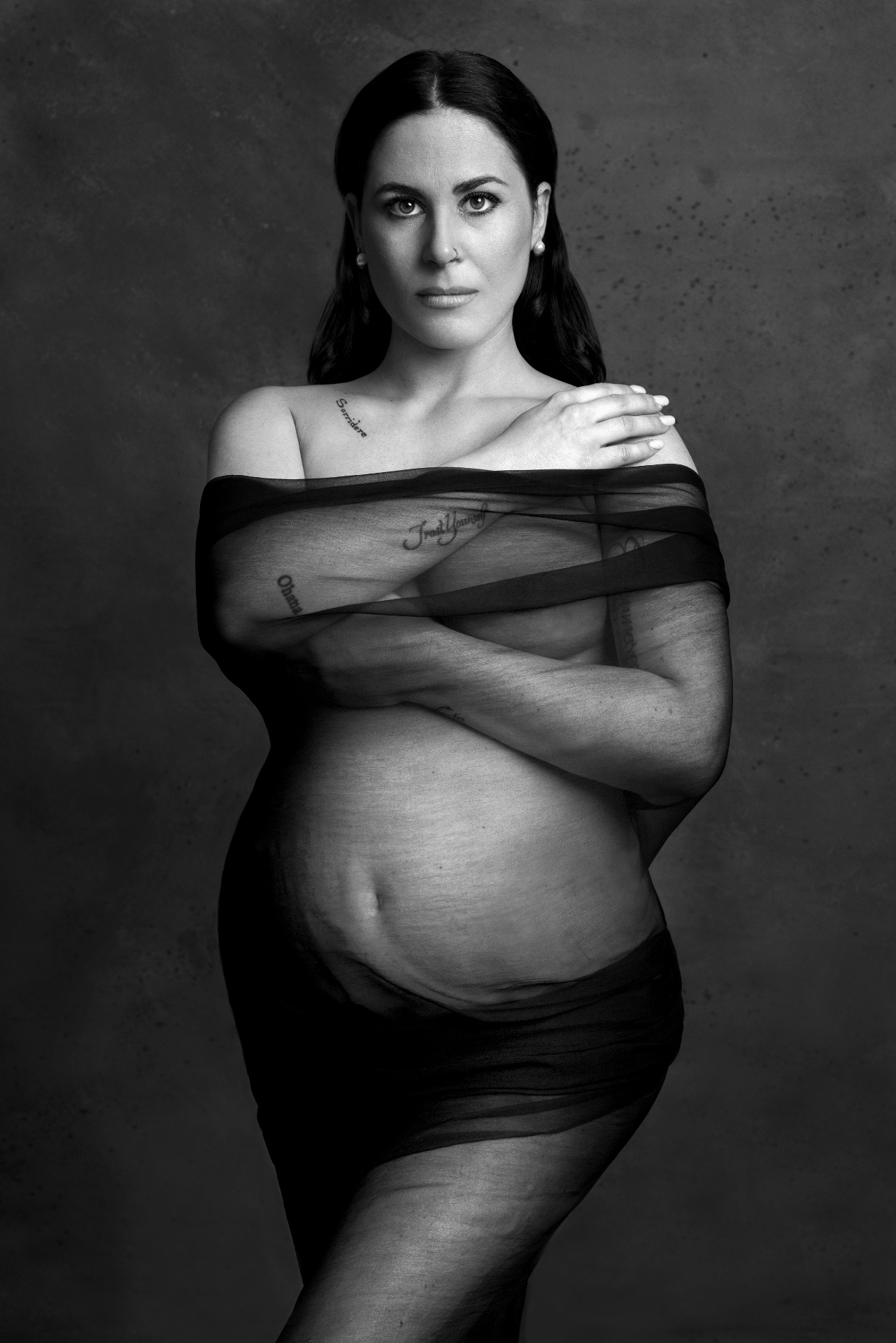 Scatto artistico in bianco e nero di maternità a Bologna realizzato da Claudia Spaziani: donna incinta con tatuaggi e tessuto trasparente, atmosfera drammatica.