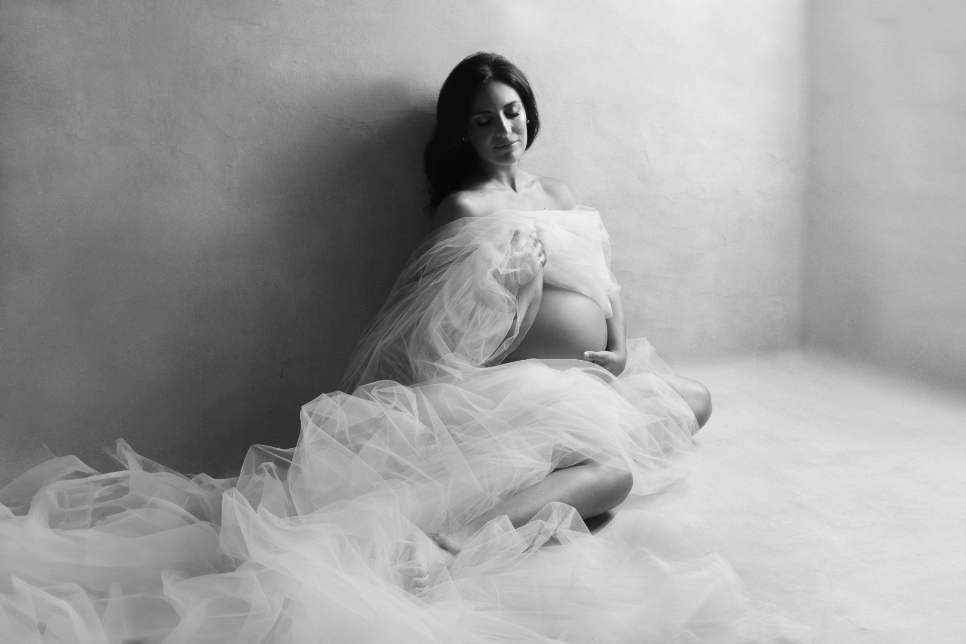 Ritratto in bianco e nero di donna incinta seduta a terra in un angolo dalle pareti chiare, avvolta in un ampio drappo di tulle. Studio fotografico Claudia Spaziani, Bologna.