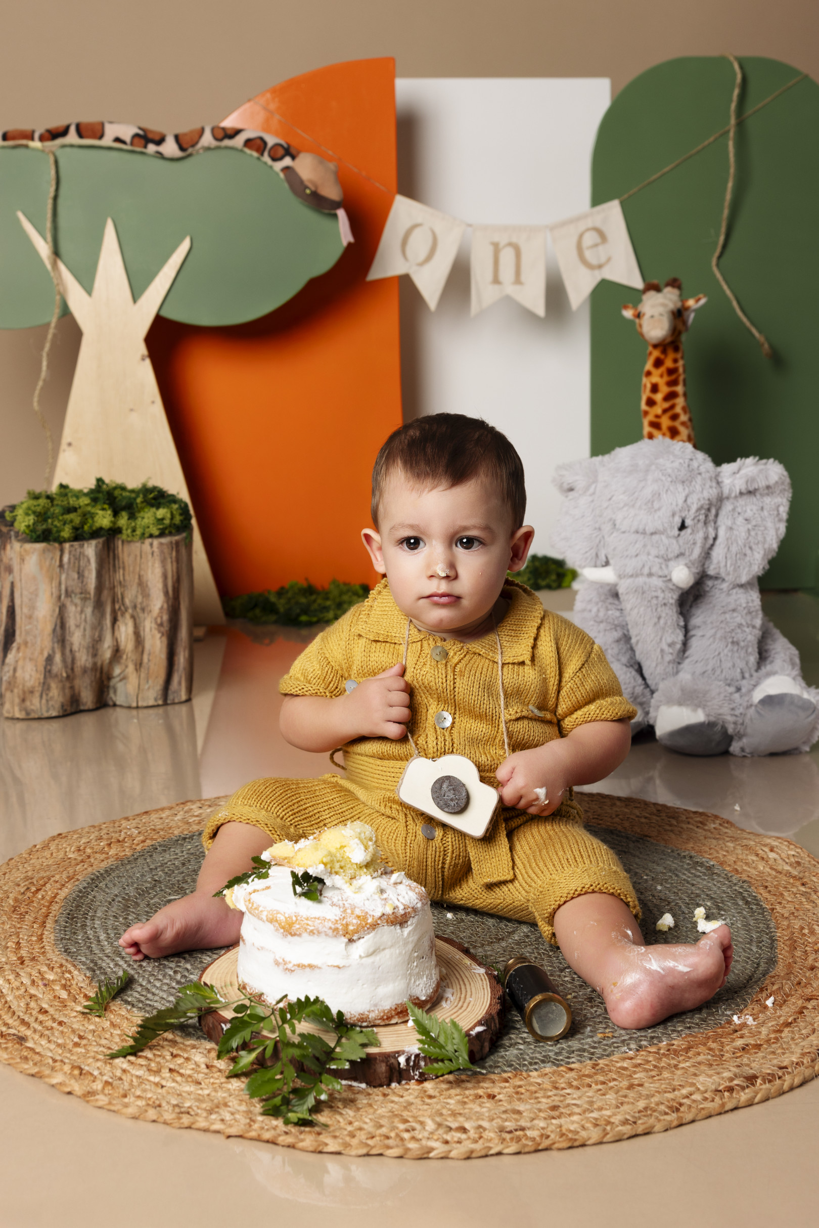 Bambino gioca con una macchina fotografica di legno accanto alla torta con felci — smash cake a tema safari, Bologna.