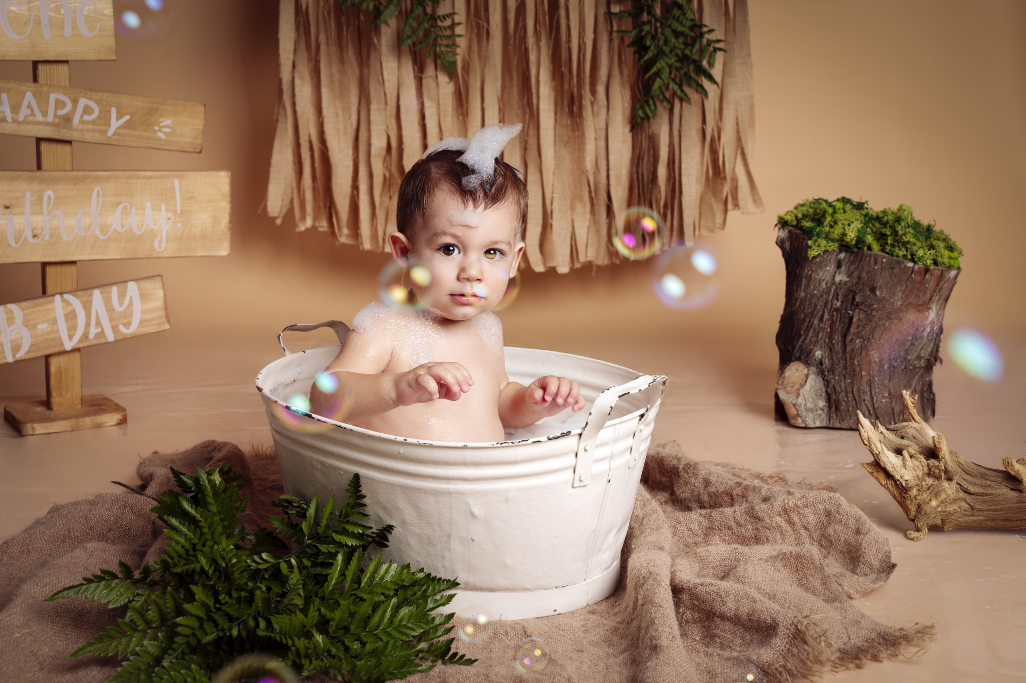Bambino in piedi nella vasca tra le bolle, sabbia e legni naturali — sessione splash boho, Bologna.