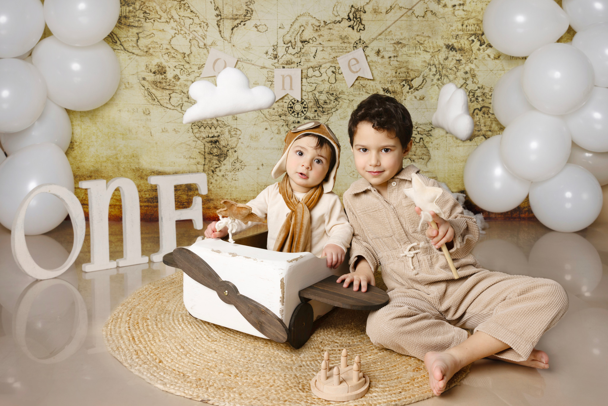 Bambino con cappellino da aviatore sull’aeroplano di legno insieme al fratello maggiore — set a tema esplorazione con lettere “ONE”, mappa e palloncini bianchi.