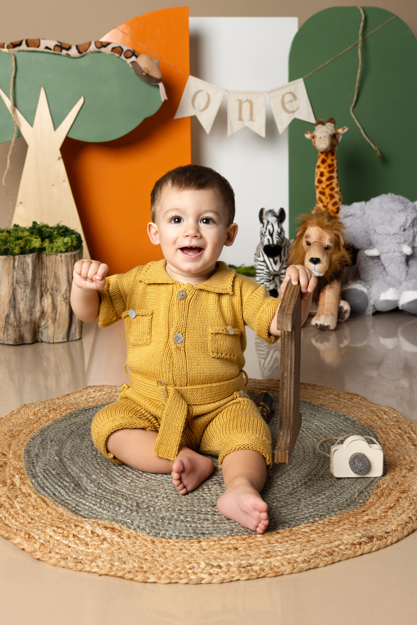 Bambino di un anno in tutina senape ride sul tappeto tenendo una lettera di legno — smash cake a tema safari a Bologna di Claudia Spaziani.