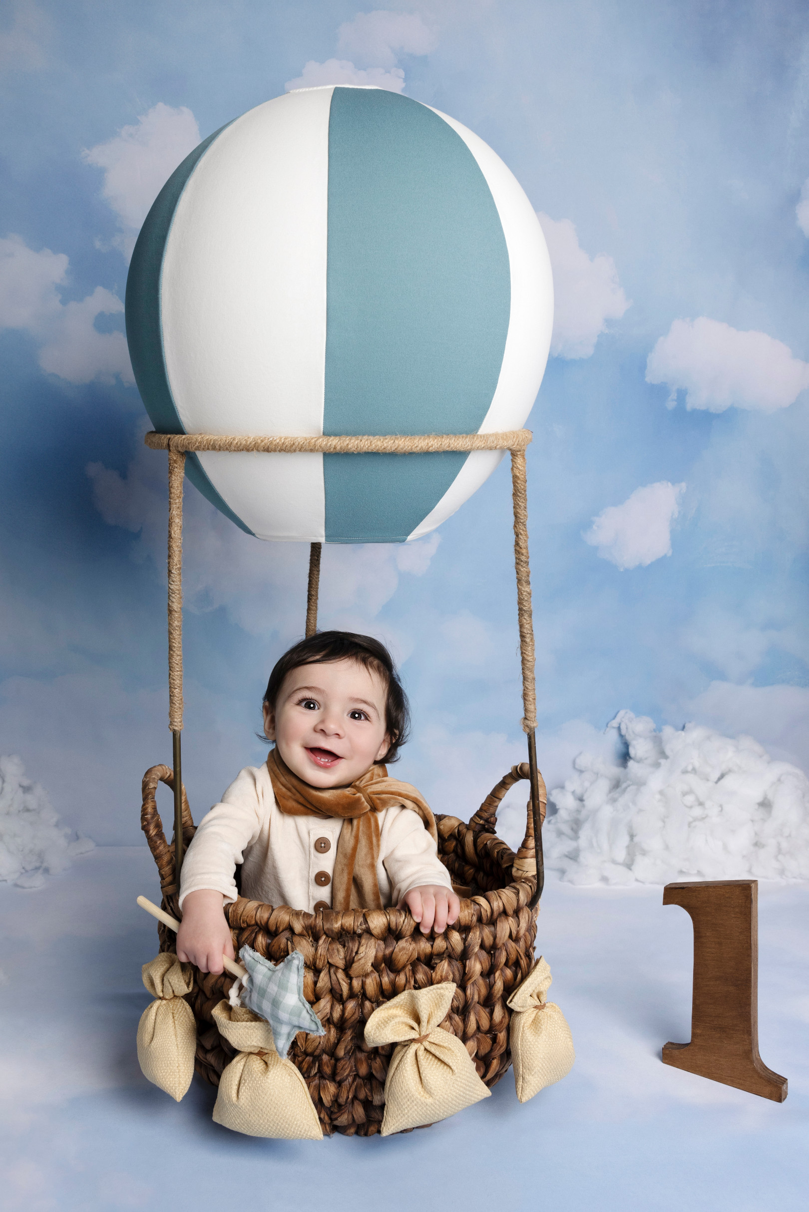 Bambino di un anno vestito da aviatore sorride nella mongolfiera di vimini, fondale cielo e nuvole, numero “1” di legno — primo compleanno a Bologna.
