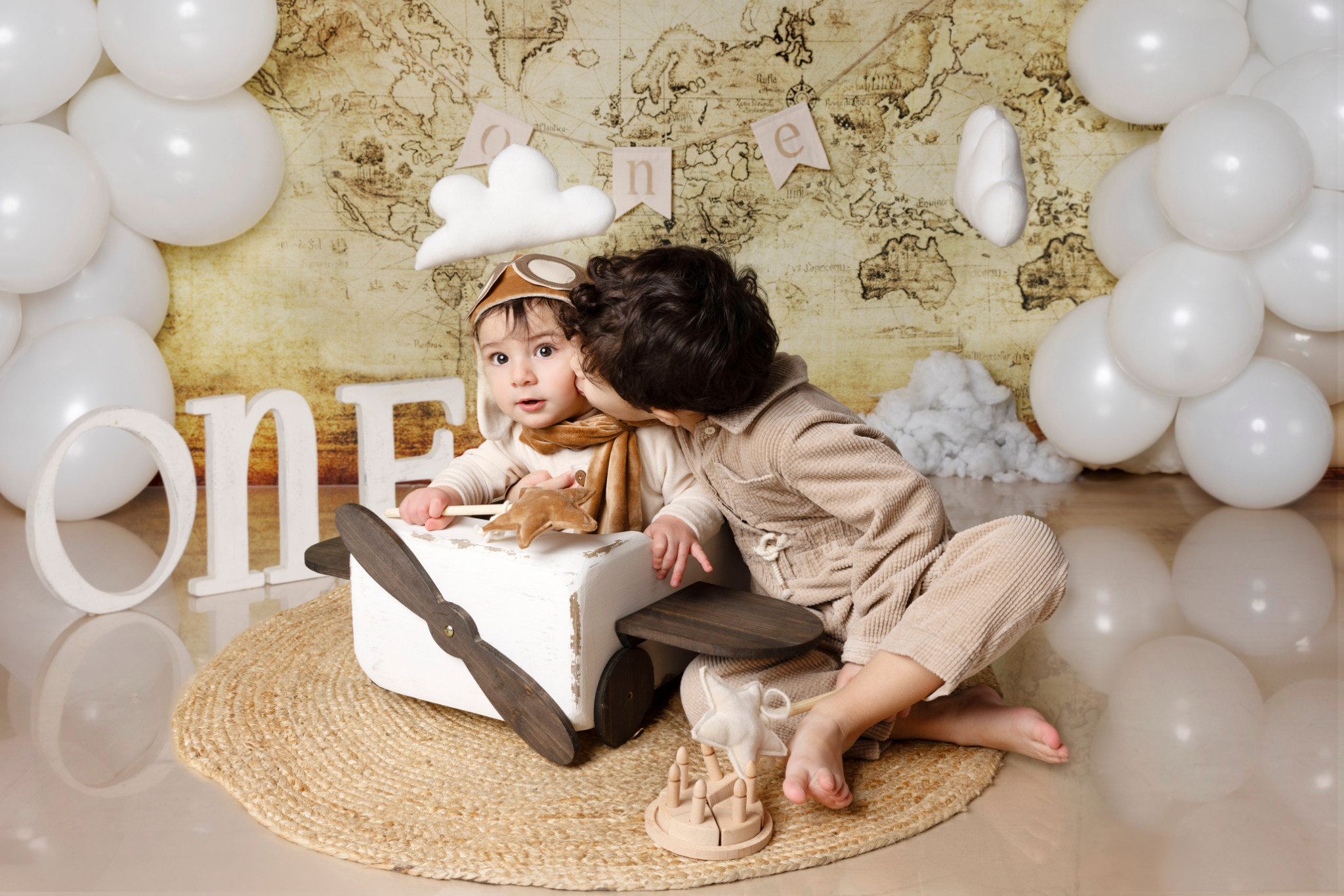 Bambino nell’aeroplanino di legno con il fratello maggiore che si sporge — set primo compleanno a tema viaggio, lettere “ONE”.