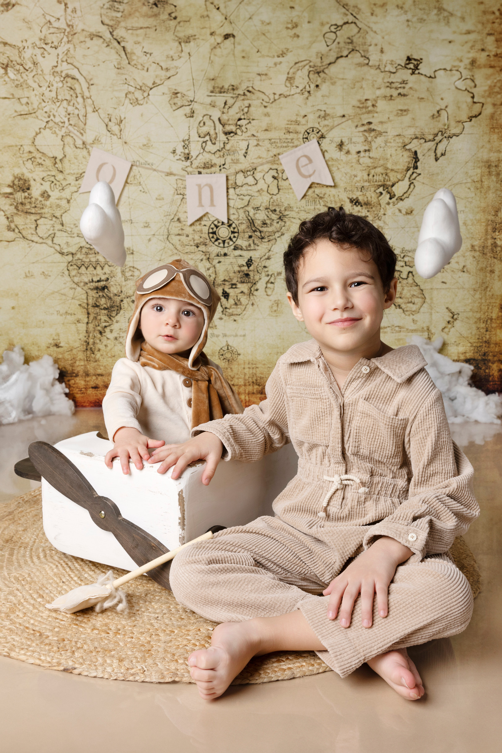 Bambino nell’aeroplanino con il fratello maggiore seduto a gambe incrociate che sorride — set a tema viaggio con mappa e palloncini.