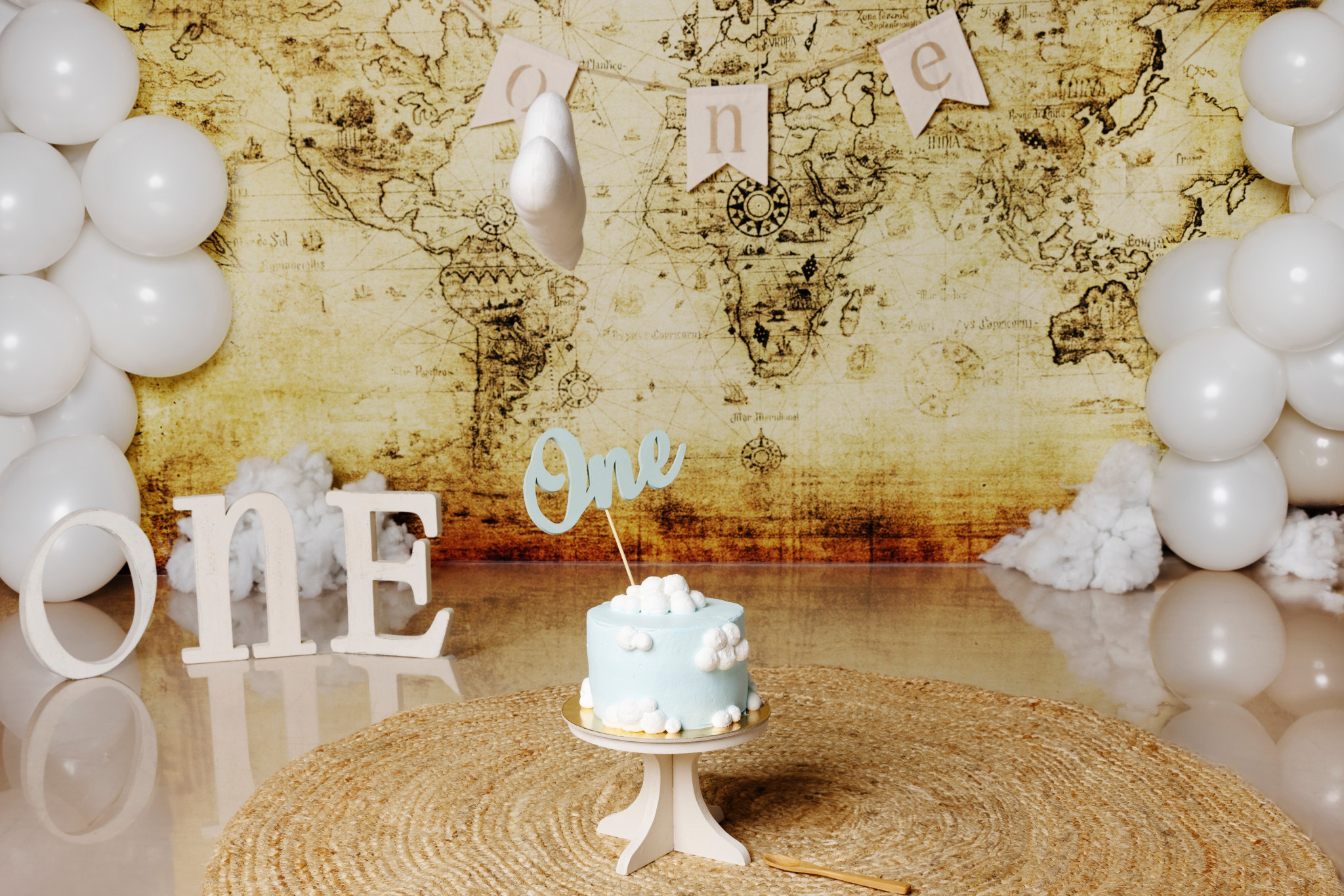 Allestimento smash cake a tema aviatore: torta azzurra con nuvole e topper “One”, lettere “ONE”, palloncini bianchi e fondale mappa antica — Bologna.