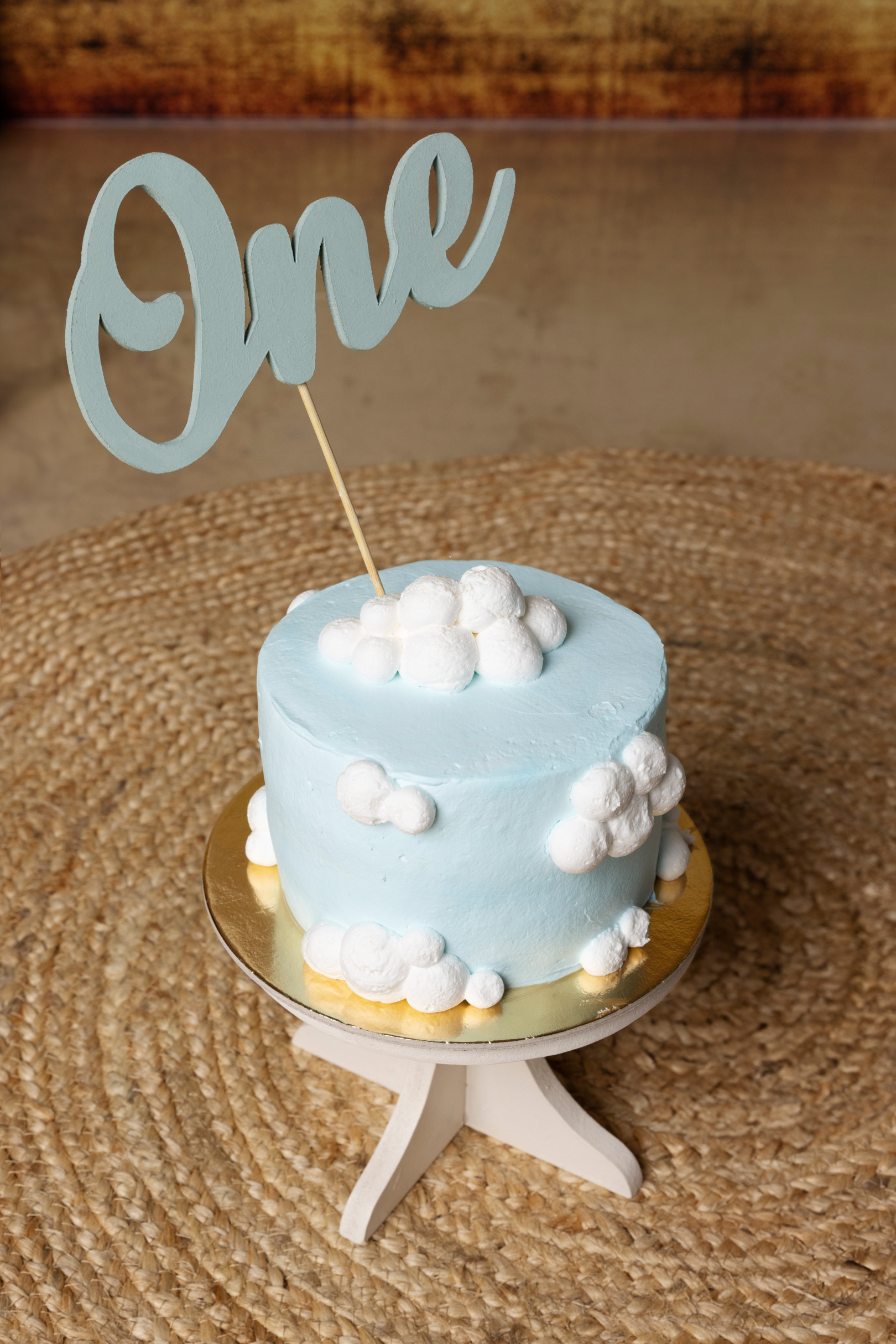 Primo piano della torta smash cake azzurra decorata con nuvole e topper “One” su base di legno — tema viaggio, Bologna.