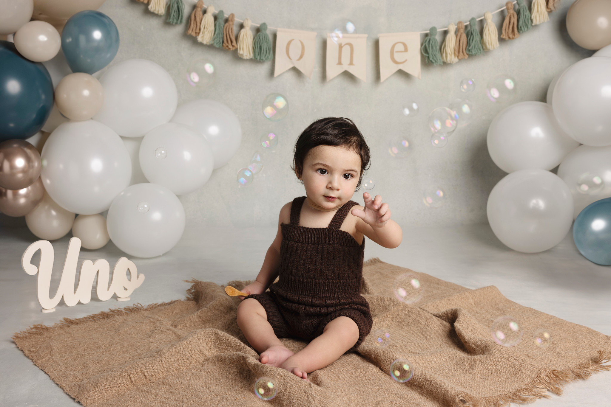 Bambino in salopette marrone su tela di juta con scritta “Uno” e ghirlanda di palloncini — primo compleanno a Bologna.