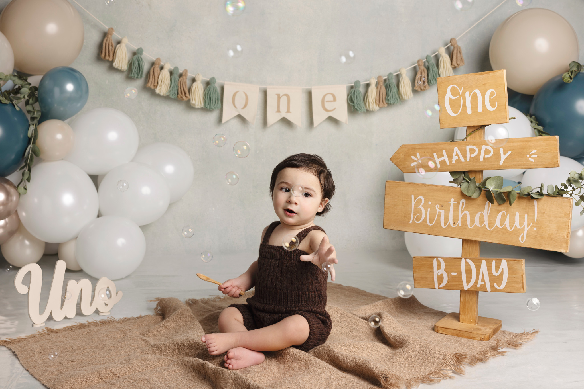 Bambino in salopette marrone con cartelli “Uno” e “Happy Birthday” e ghirlanda di palloncini — set primo compleanno a Bologna.