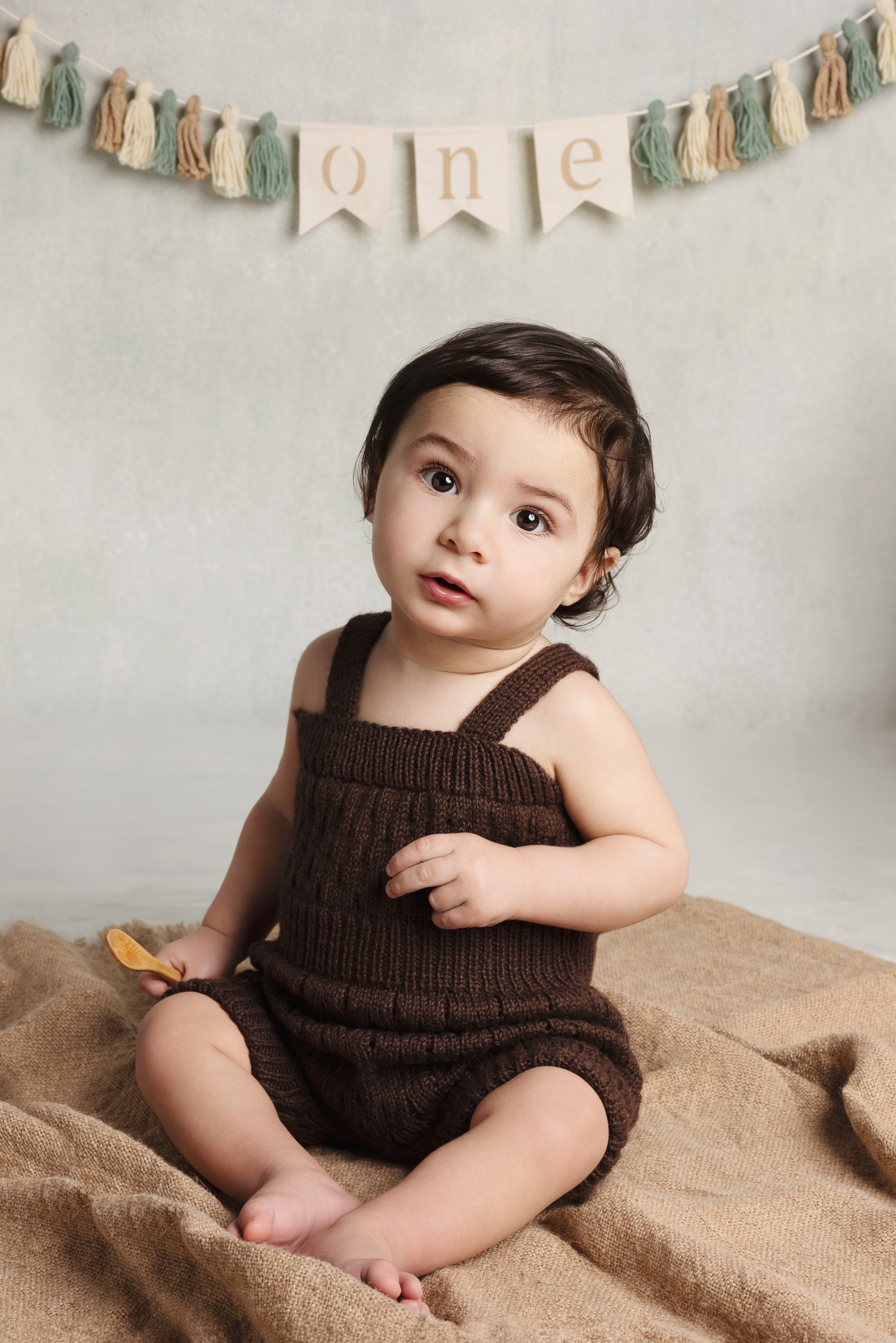 Bambino di un anno in salopette di maglia marrone seduto con un biscotto — fondale neutro con festone “one”, Bologna.