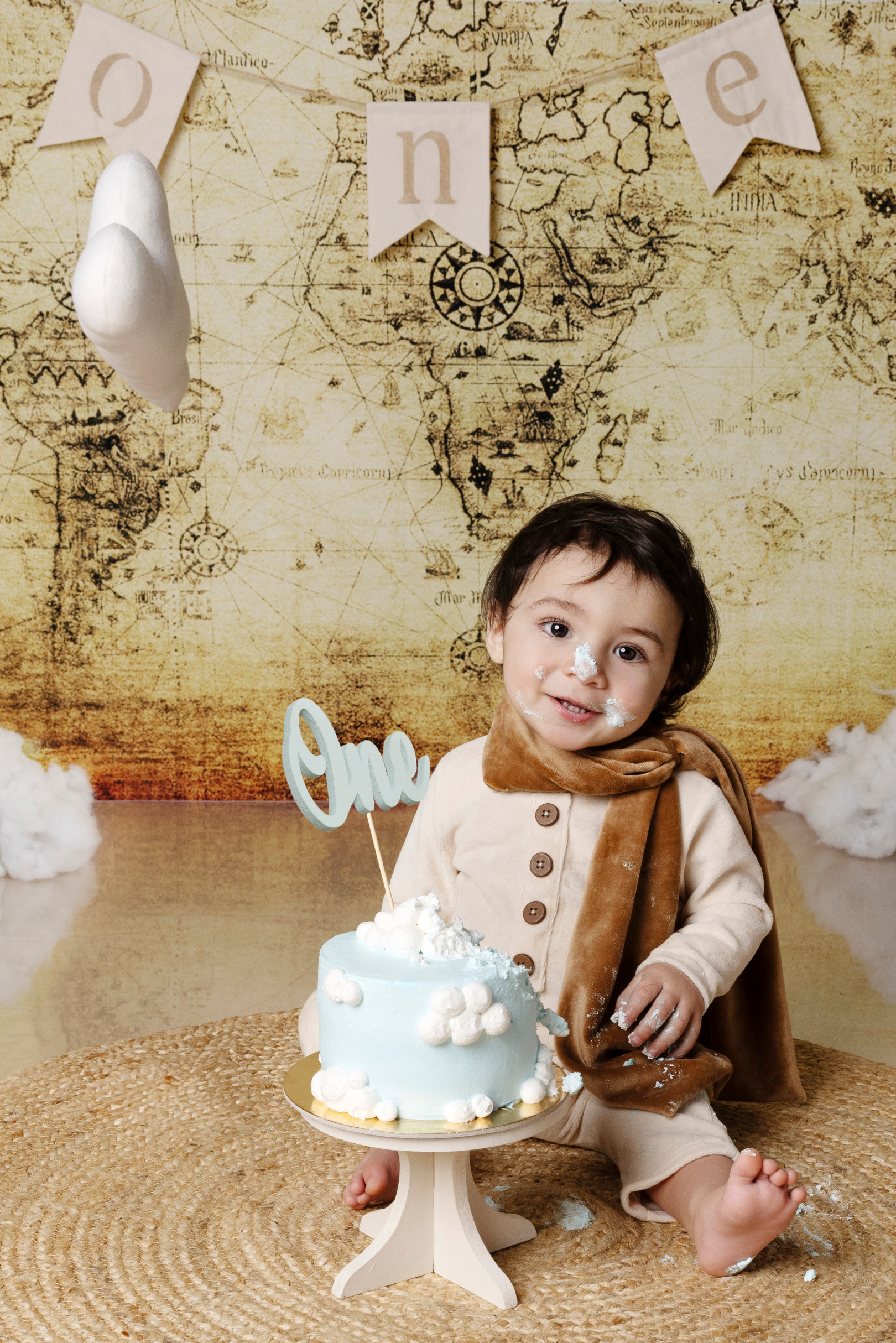 Bambino con sciarpa allunga la mano verso la torta azzurra con topper “One” — smash cake a tema viaggio, Bologna.
