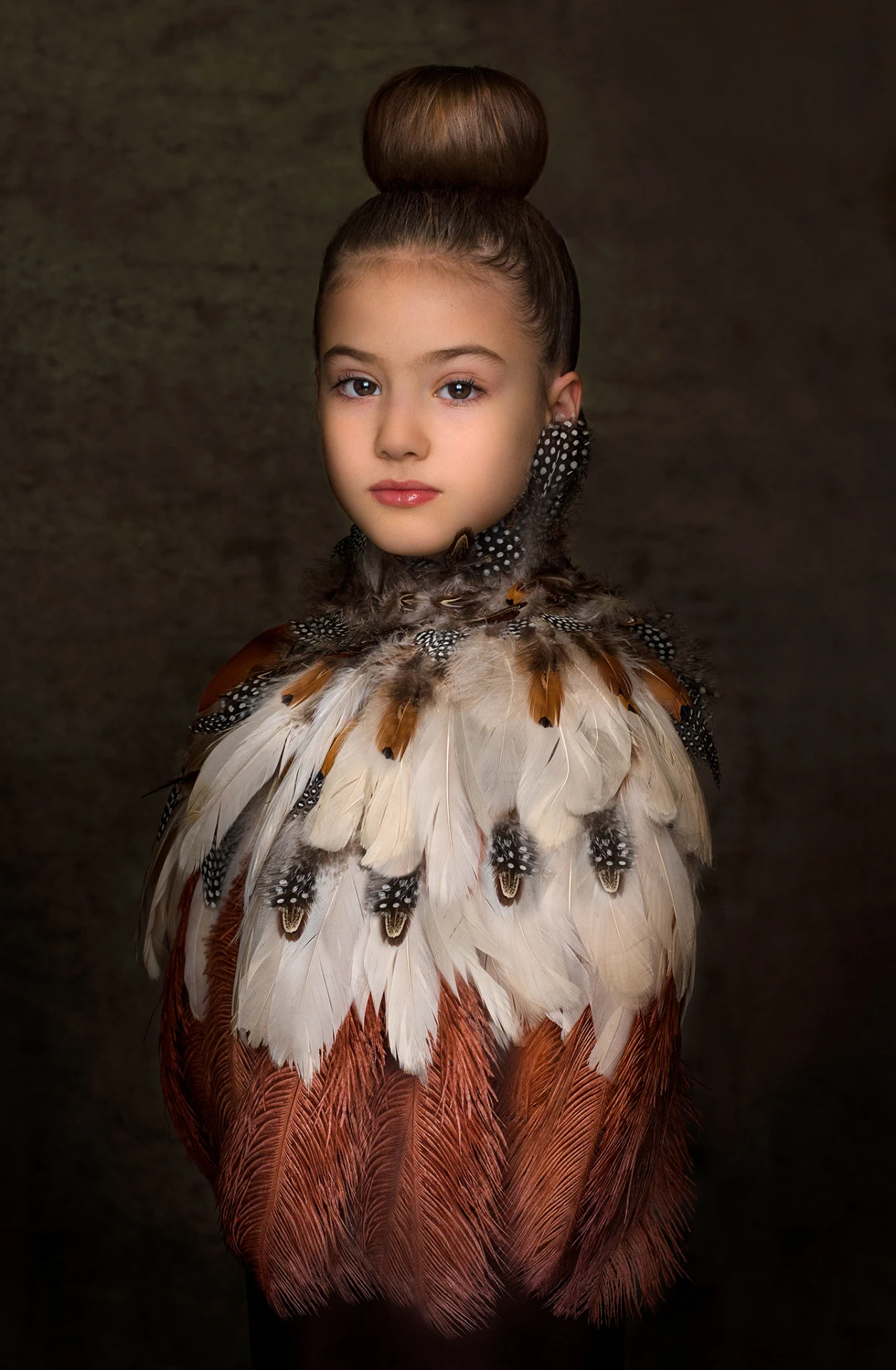 Ritratto artistico di bambina con abito di piume multicolore e chignon alto su sfondo scuro, ritratto fine art a Bologna