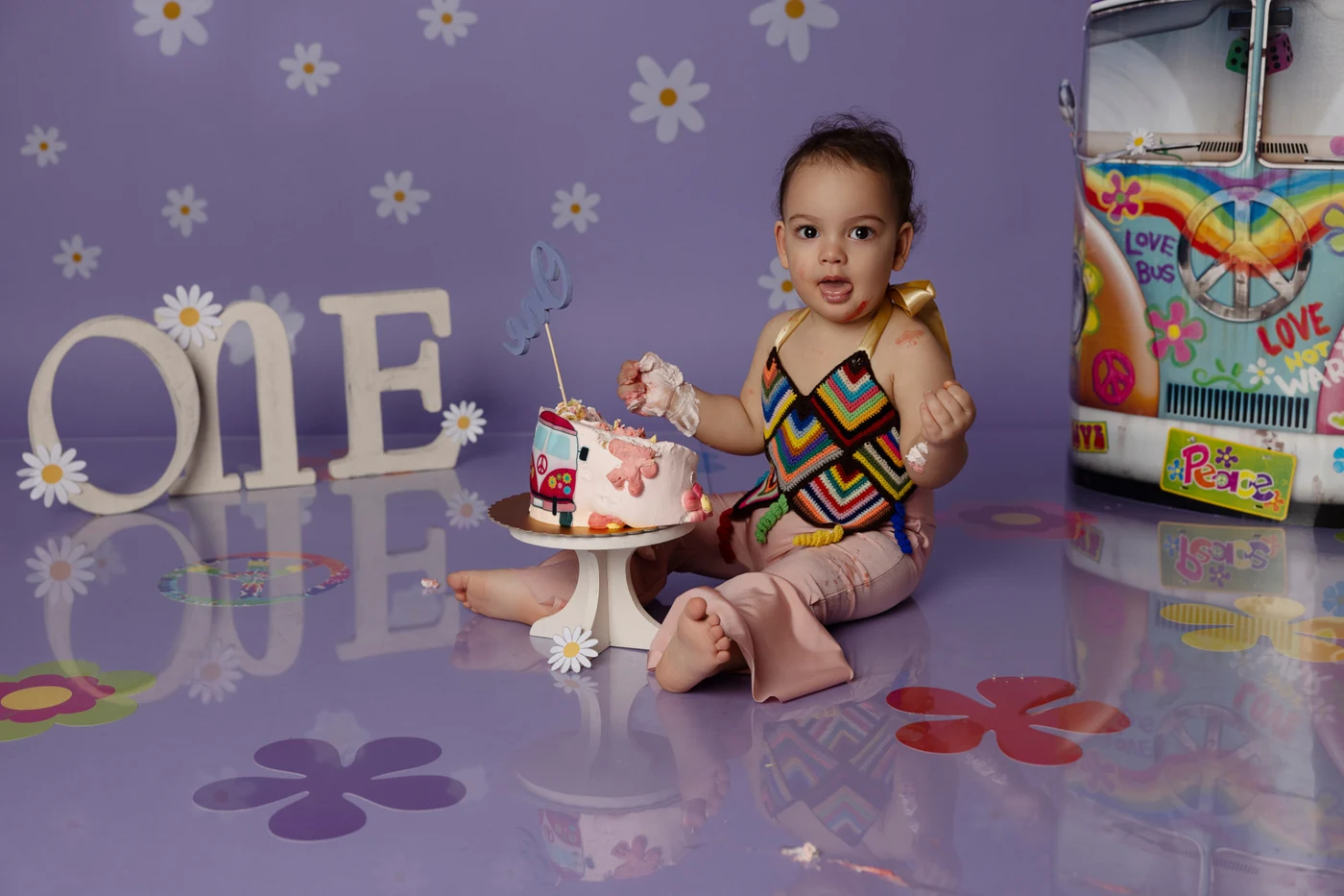 Bambina con mani sporche di torta davanti a una torta decorata con lettere "ONE", pavimento viola — smash cake primo compleanno a Bologna.