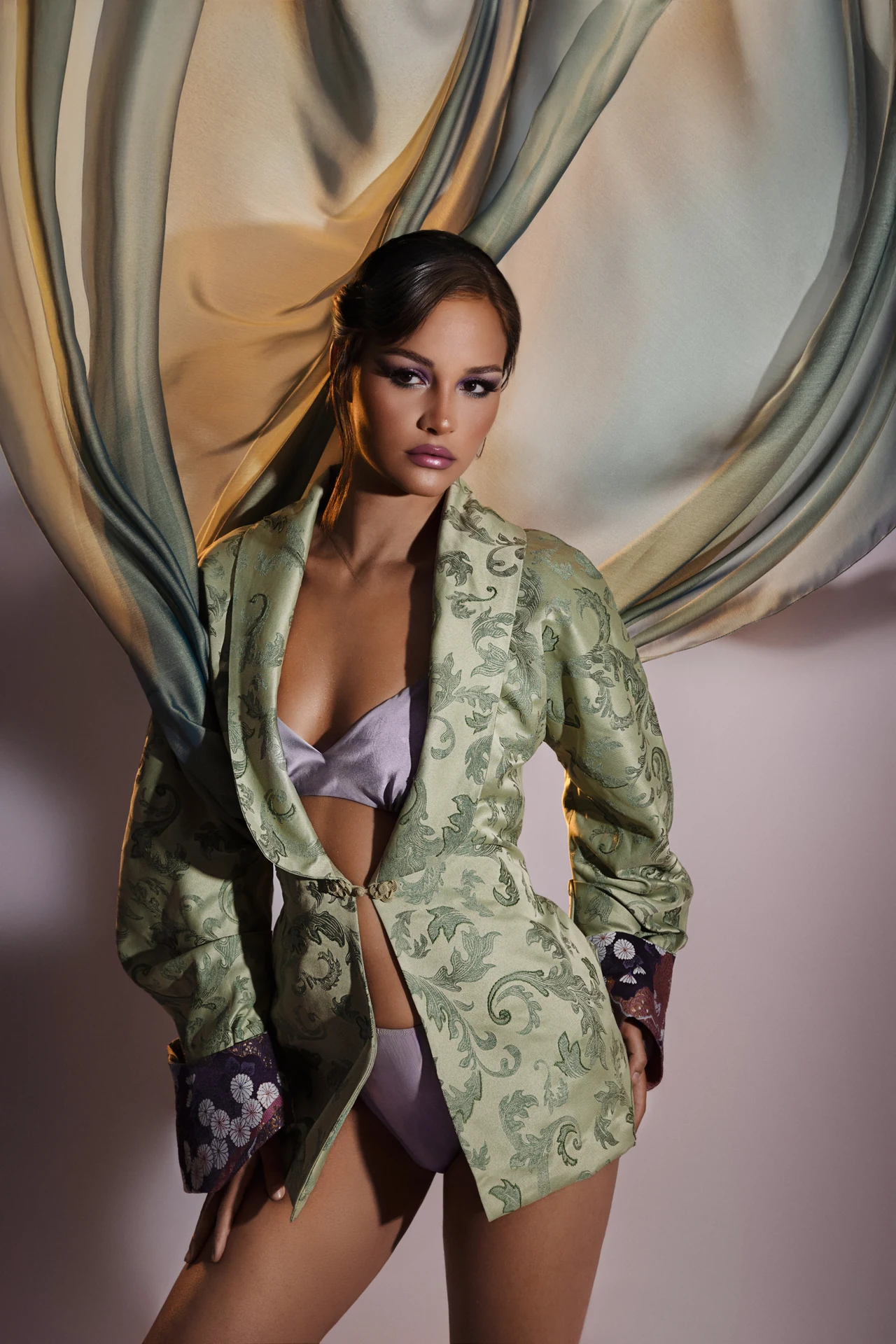 Ritratto glamour fine art a Bologna: modella in giacca di broccato verde e lingerie lavanda, su fondale di tessuto fluttuante.