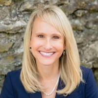 Smiling woman with straight blonde hair wearing a dark blazer and pearl necklace, in front of a stone wall.