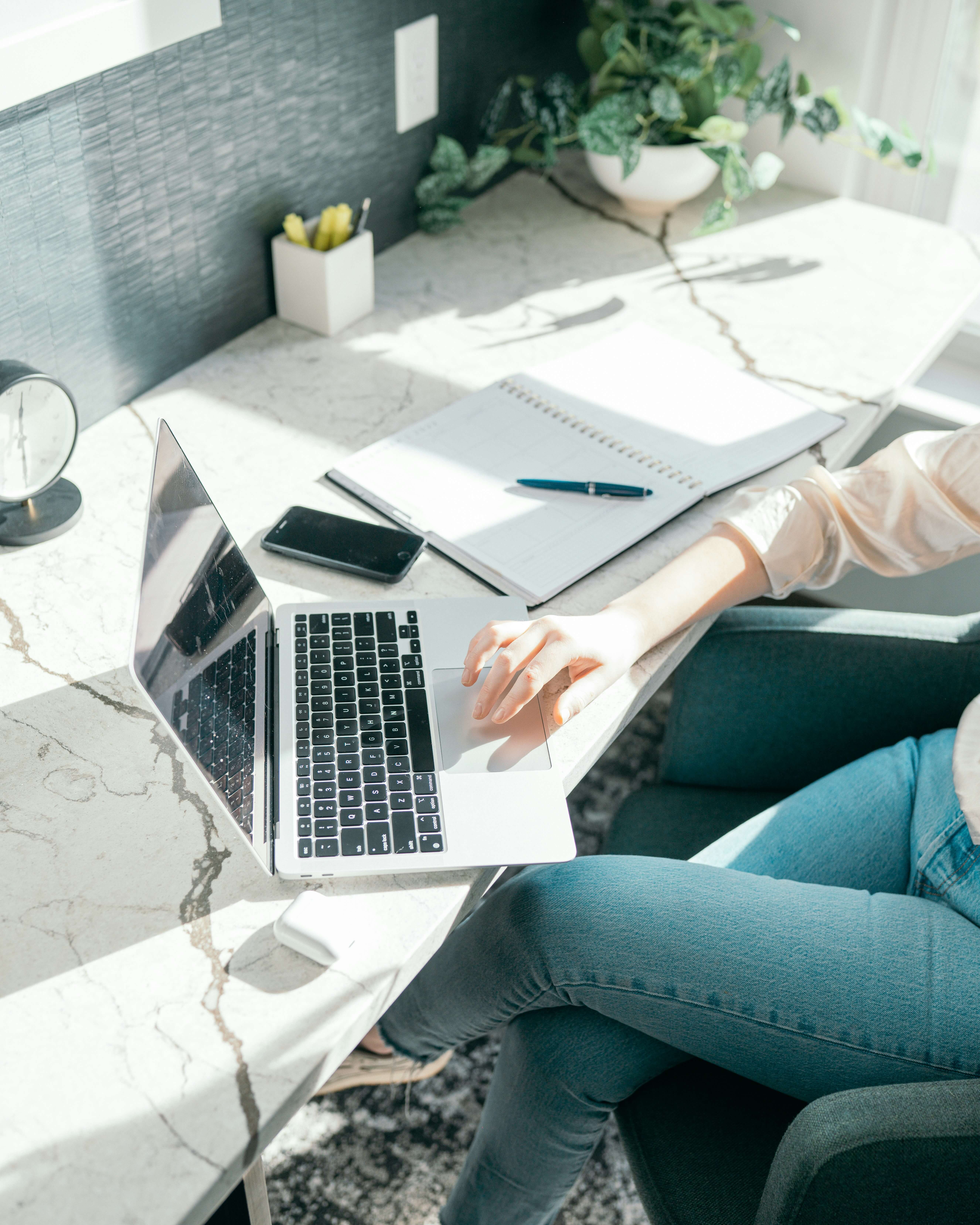 Person sitting at a marble desk with crossed legs using a laptop, with a smartphone, notebook, pen, clock, and potted plant also on the desk in bright natural light.