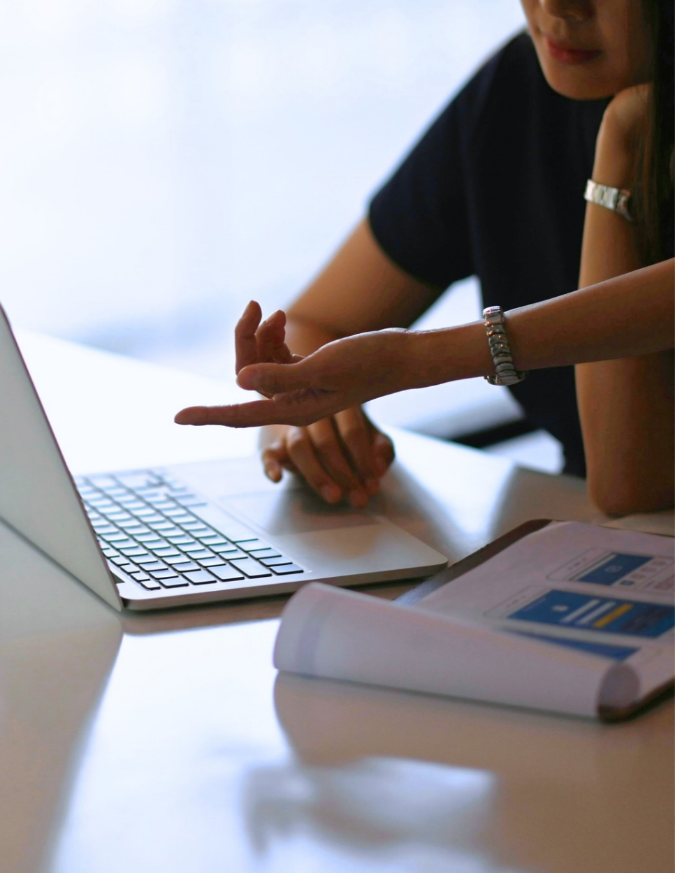 Person sitting at a table using a laptop with one hand on the keyboard and the other hand gesturing, with a rolled-up document showing mobile app design layouts beside the laptop.