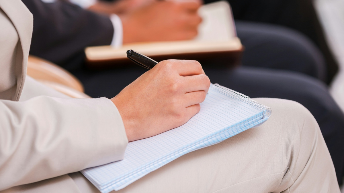 Person wearing a light-colored suit sitting and writing on a spiral-bound notebook with grid paper using a black pen.