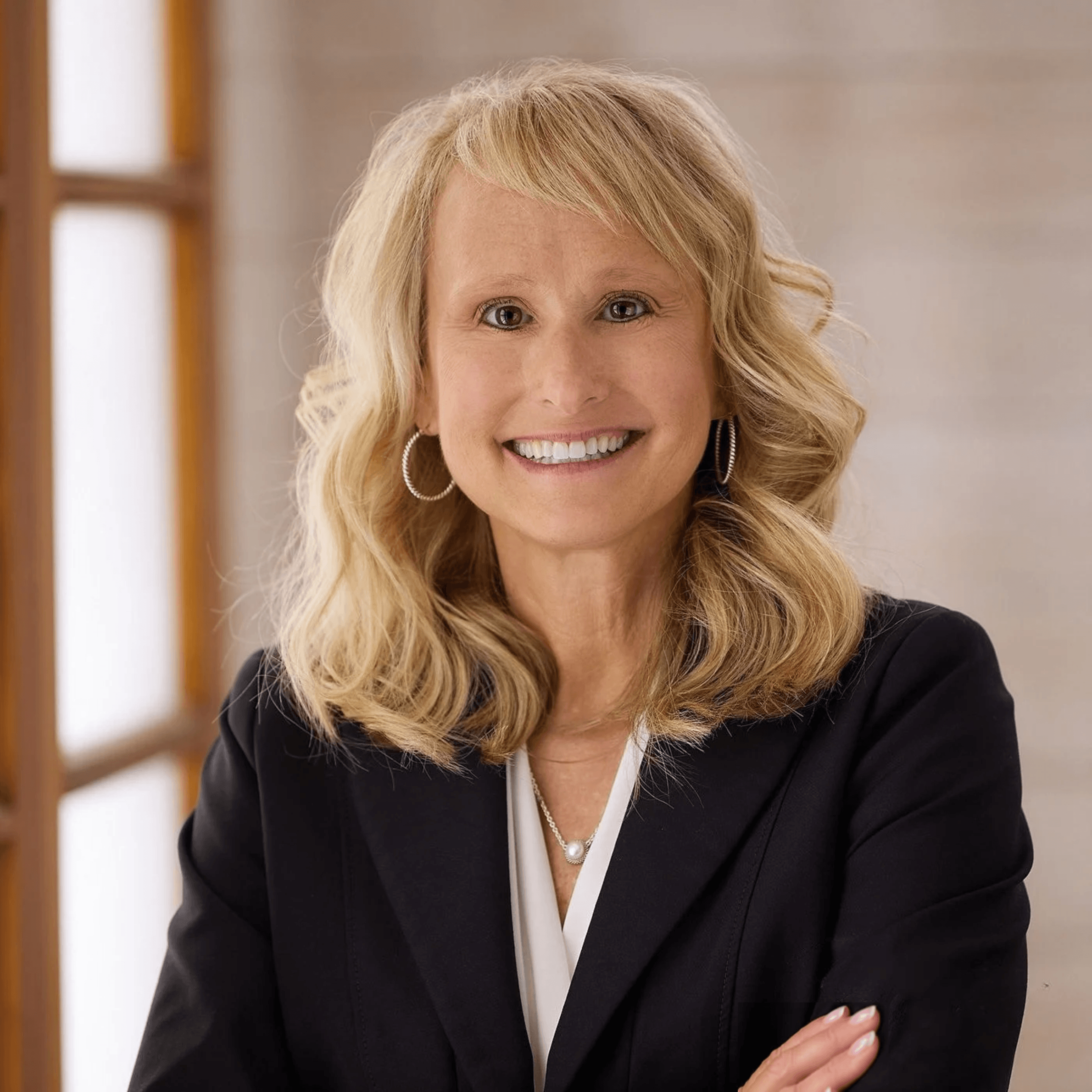 Smiling woman with blonde wavy hair wearing a black blazer, white blouse, pearl necklace, and hoop earrings against a softly lit background.